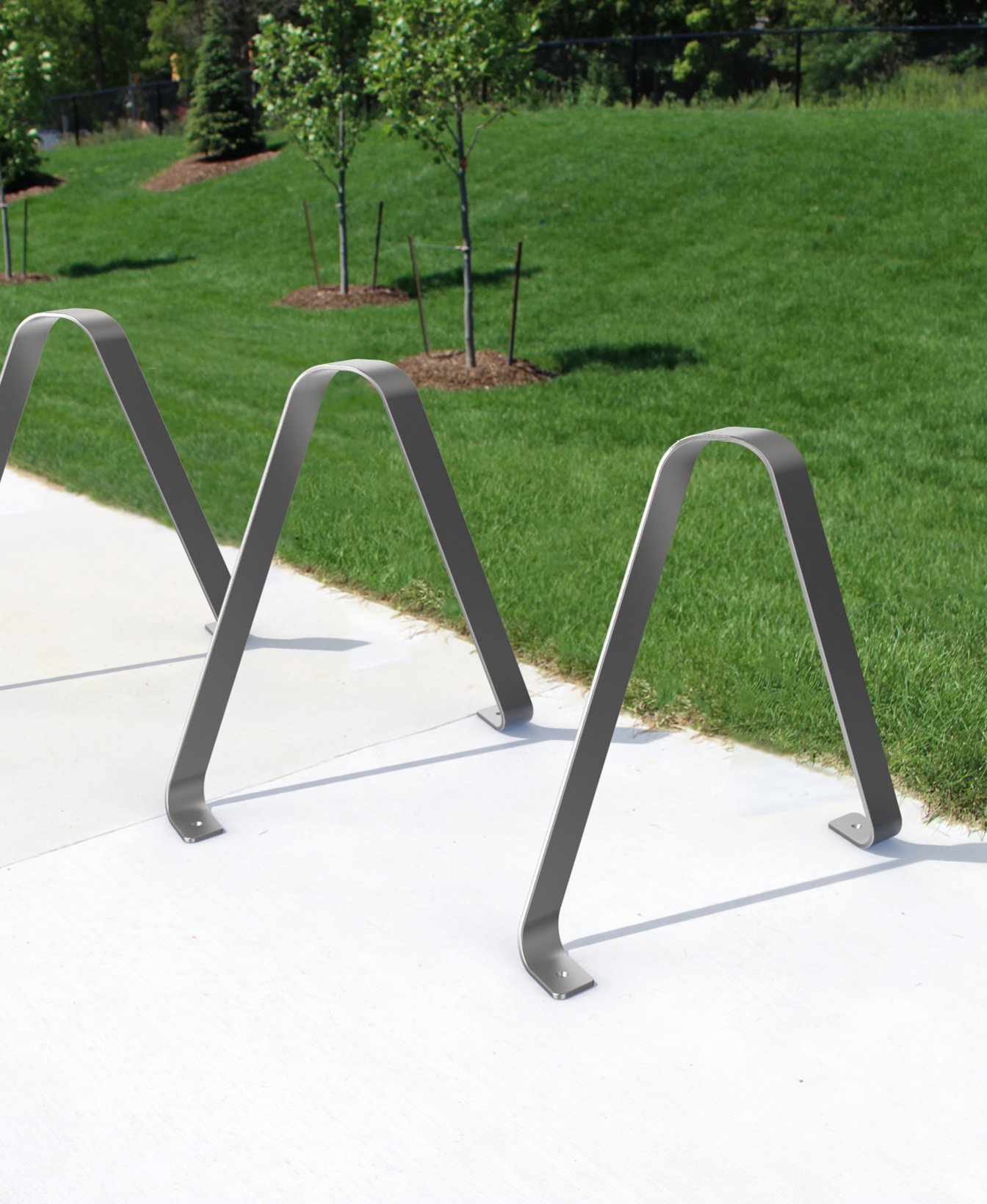 3100 Bike Racks, shaped like inverted U’s, are installed on a concrete sidewalk beside a grassy area with young trees.