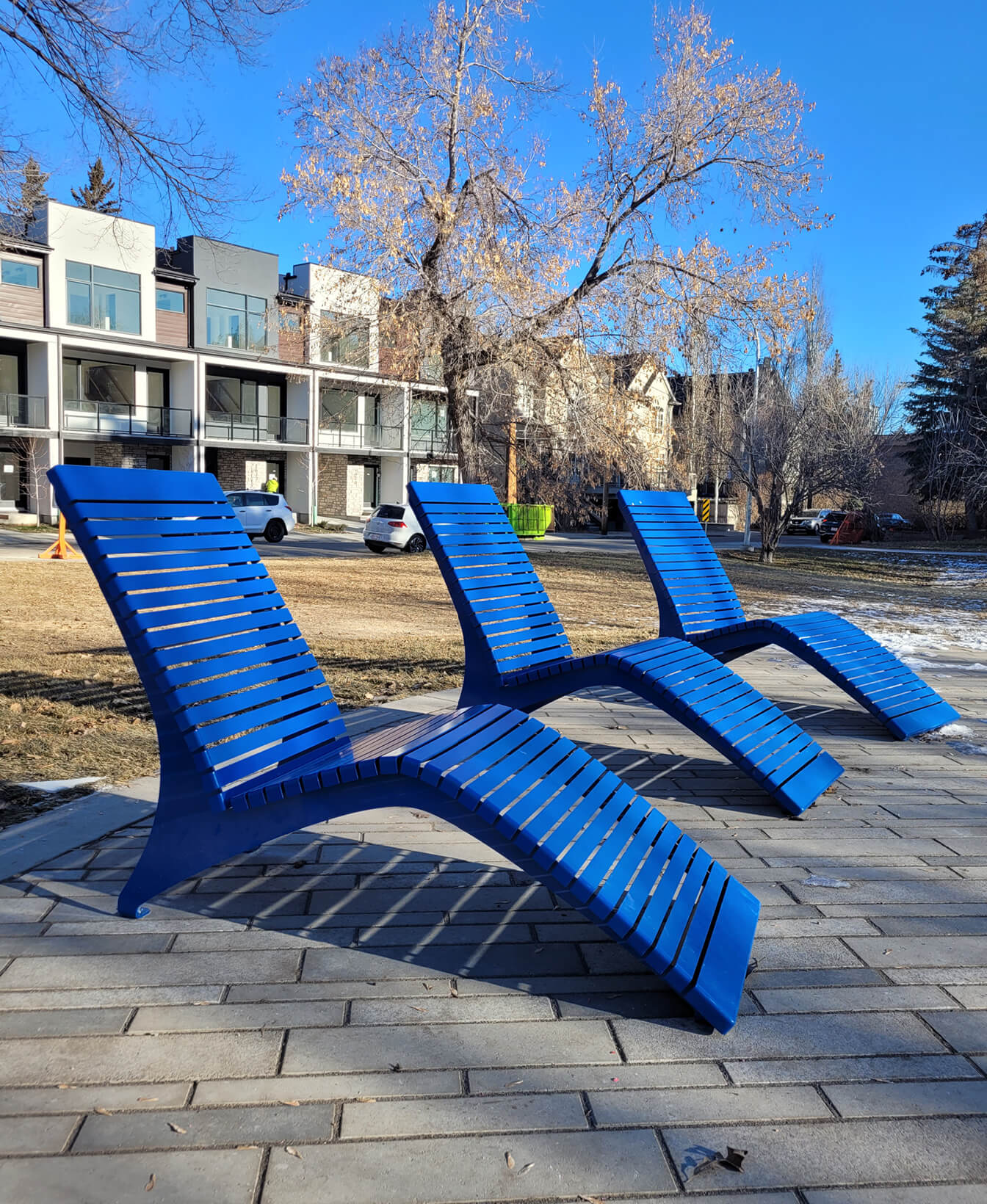 Three 720 Chaise Lounge chairs in blue are set on a paved area in a park, with leafless trees and residential buildings visible beneath a clear blue sky.