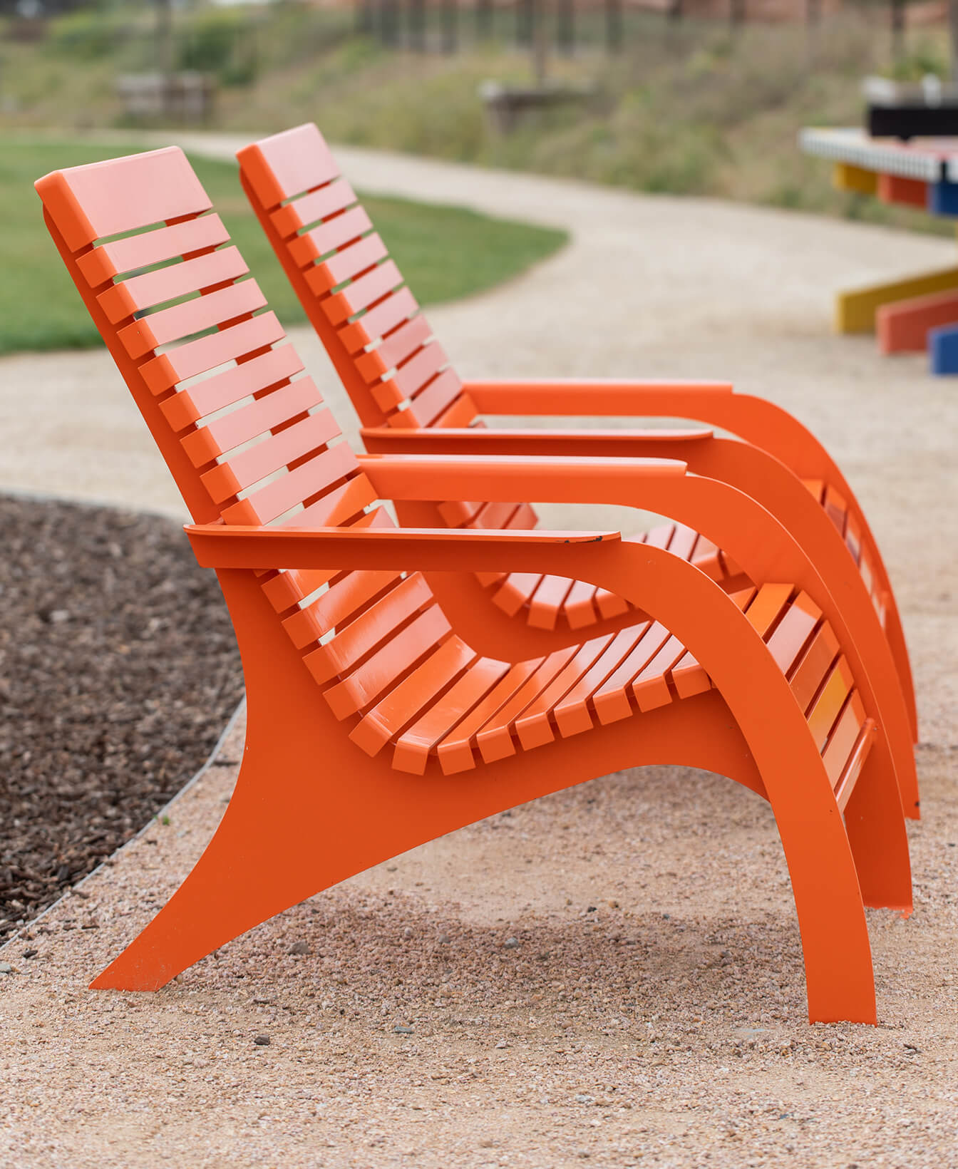 Two 720 Chairs with armrests and modern orange slats are placed on a gravel path in an outdoor park setting.