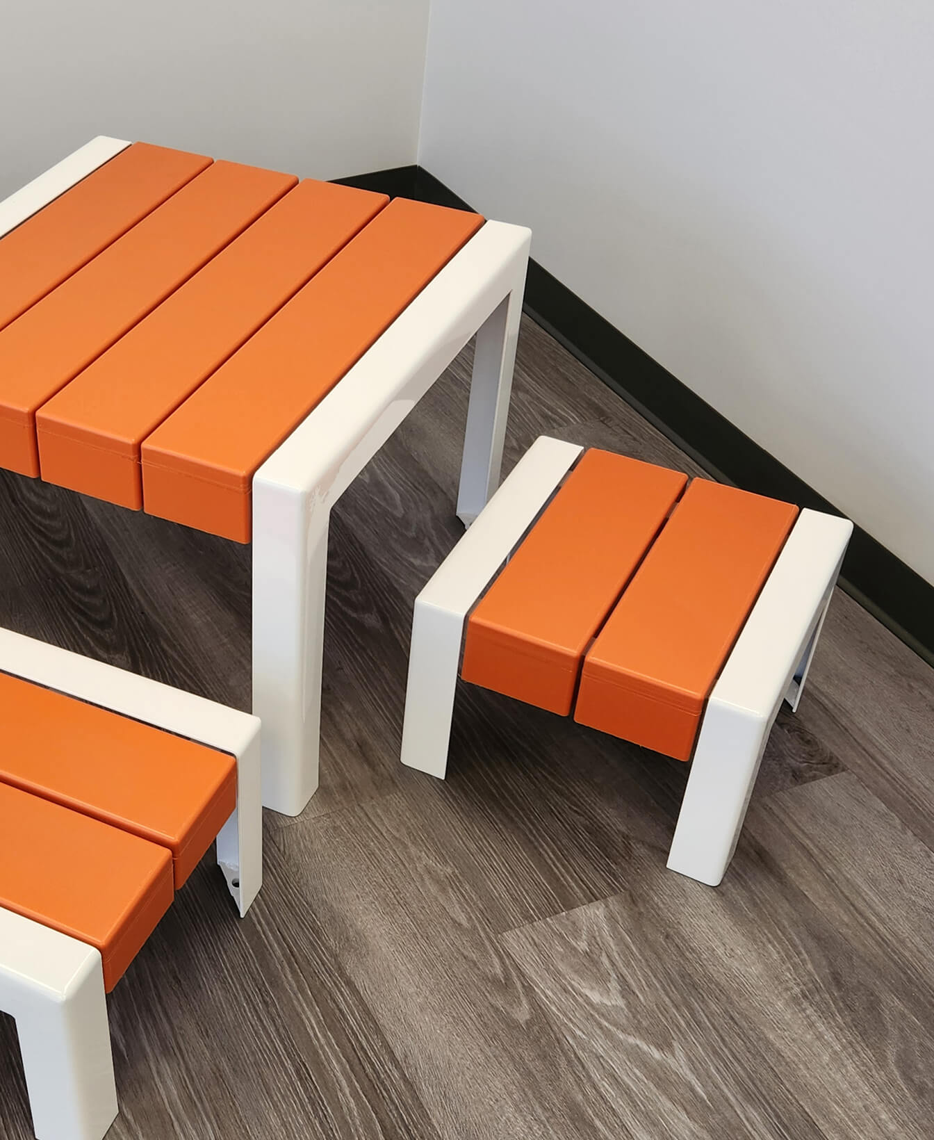 Le Tabouret 1050 pour enfants, une petite table avec deux tabourets assortis dans des cadres blancs et des lattes orange, est posé sur un sol en bois à côté d'un mur blanc.