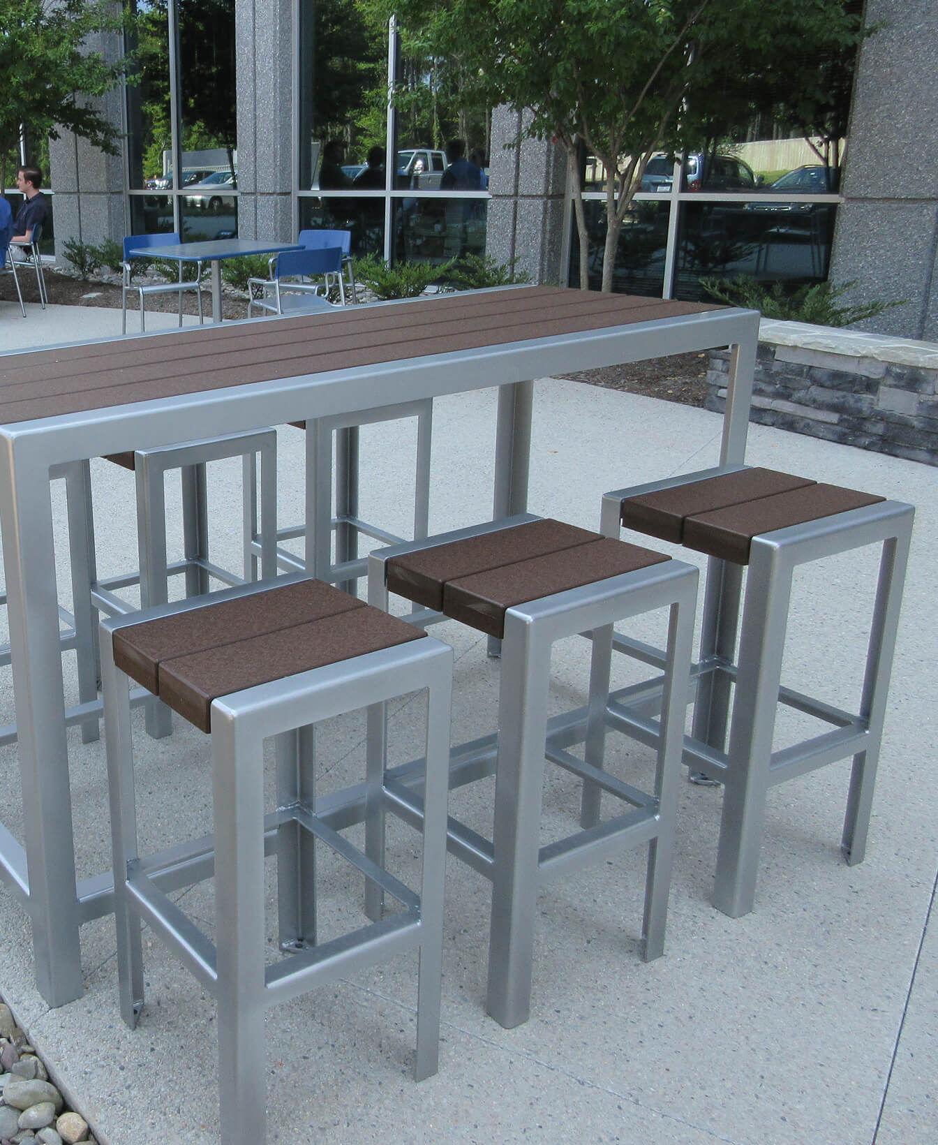 Outdoor patio featuring a tall metal table and four 1050 Bar Height Stools with brown wooden seats, set on a concrete surface beside a modern building.