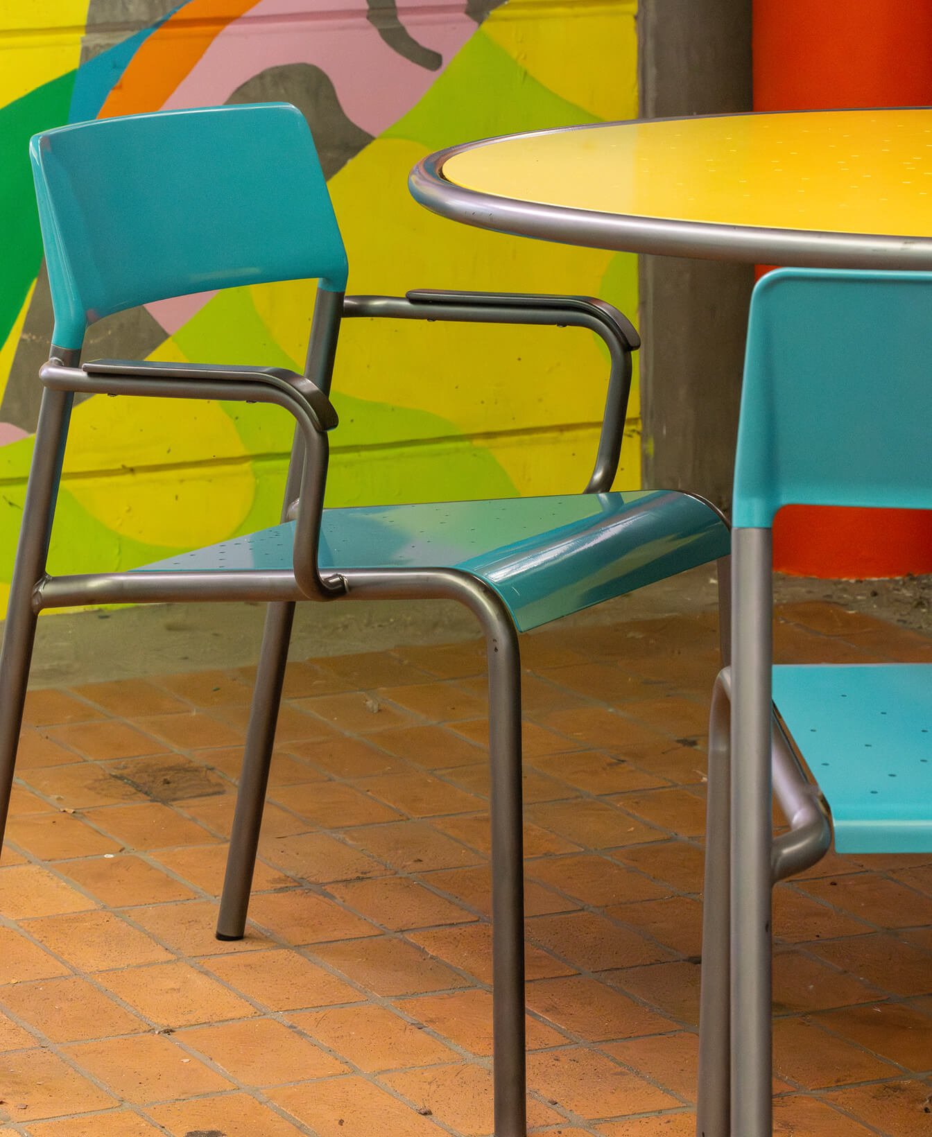 Two Foro Café Chairs in blue metal with a round yellow table on a tiled floor, set against a colorful painted wall.