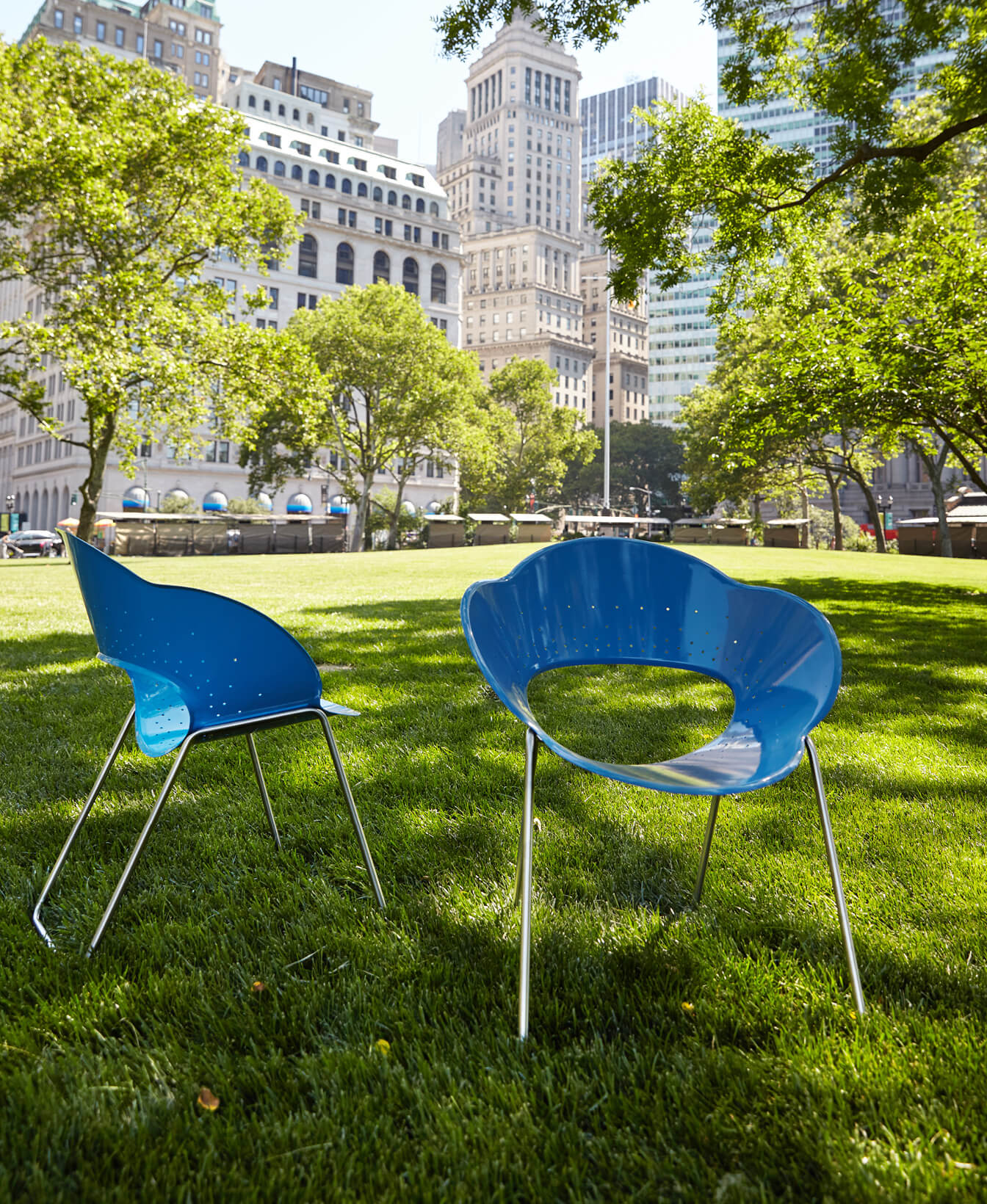 Two Battery Chairs with blue seats and metal legs are set on grass in a city park, framed by tall buildings and green trees.