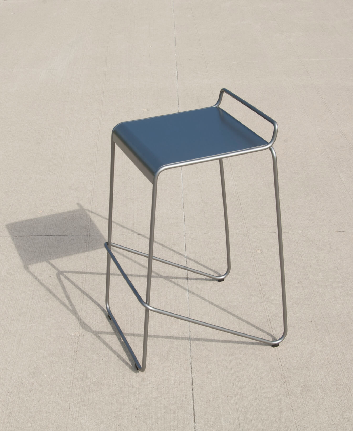 The Kontur Bar Height Stool, featuring a blue seat, metal frame, and footrest, sits on a light concrete surface and casts a shadow to the left.