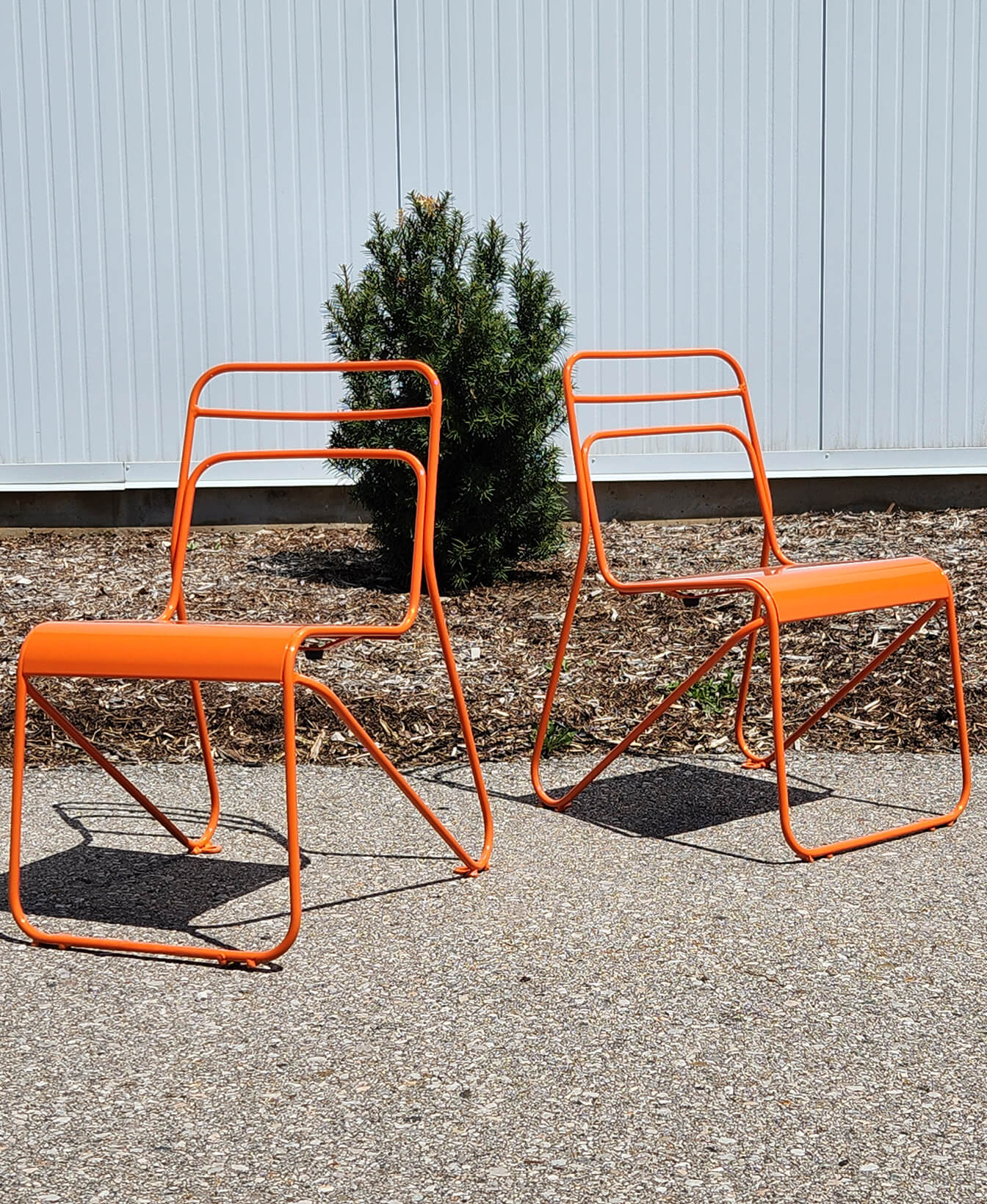 Deux chaises Mini chaise Kontur en métal orange sont assises sur une surface extérieure pavée, avec un petit arbuste à feuilles persistantes et un mur en métal à l'arrière-plan.