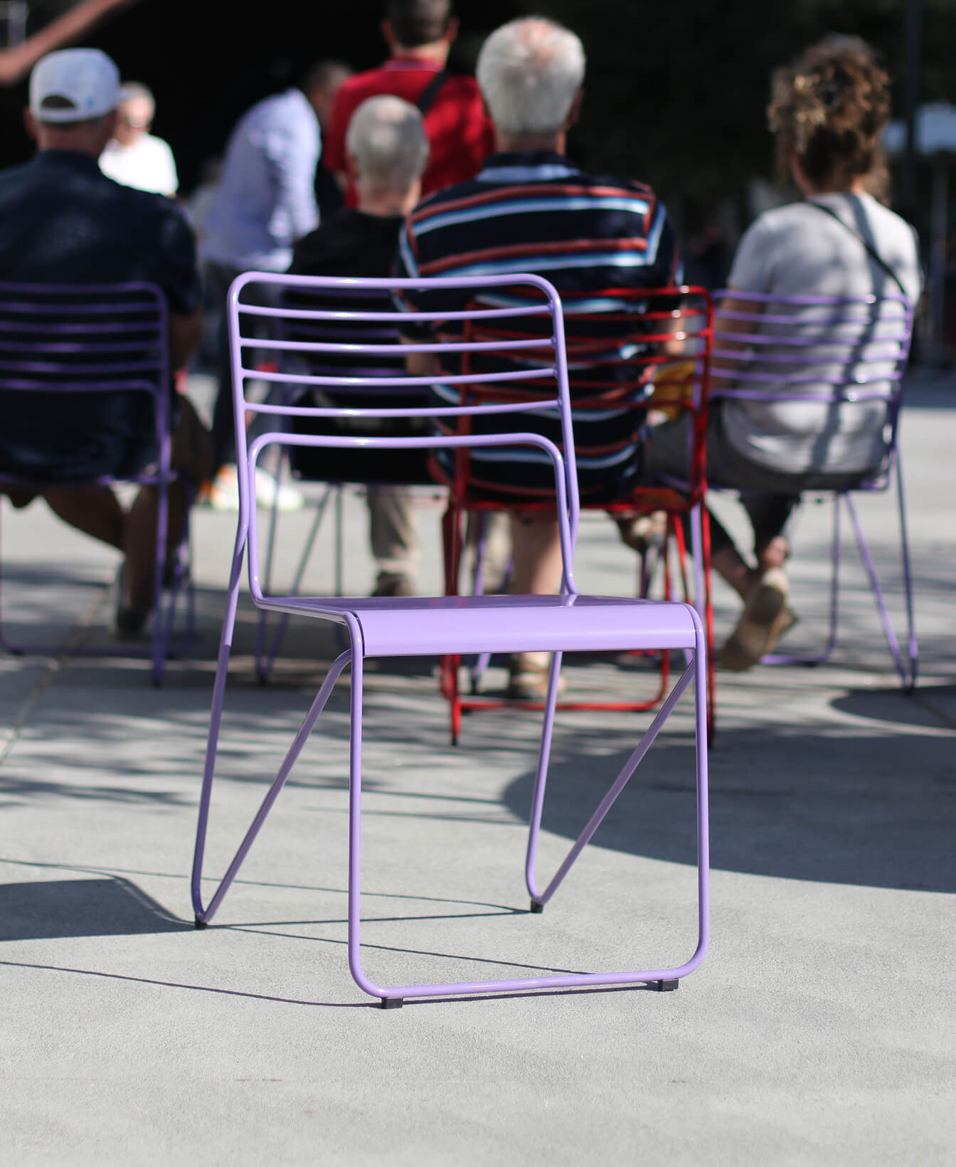 A Kontur Café Chair in light purple is in focus on a sunny outdoor plaza, with several people sitting on similar Kontur Café Chairs in the background.