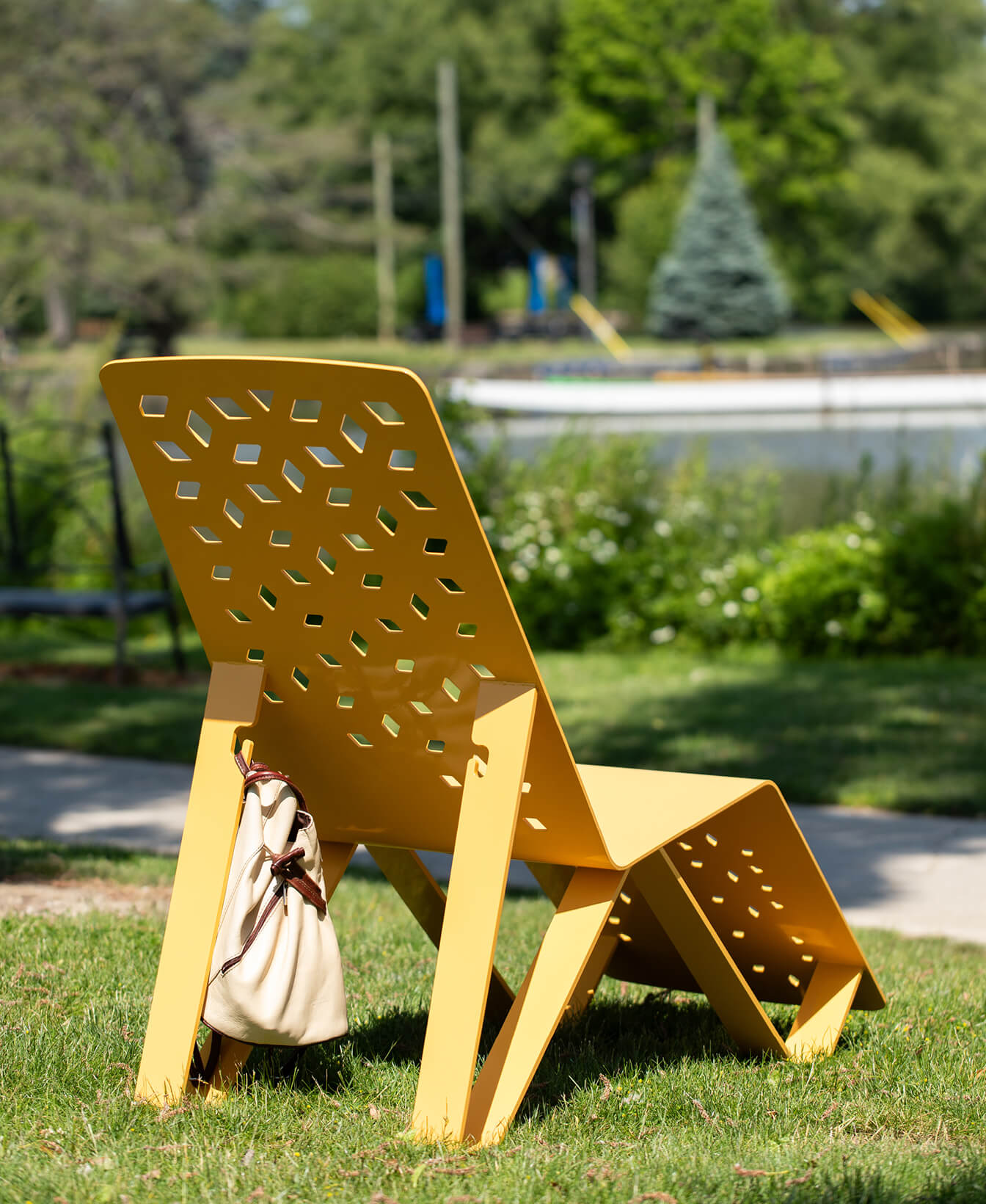 Une chaise longue en ALUM jaune, avec des découpes hexagonales, est posée sur de l'herbe. Un sac à cordon beige est suspendu à l'un de ses pieds. Des arbres et un étang flou sont visibles à l'arrière-plan.