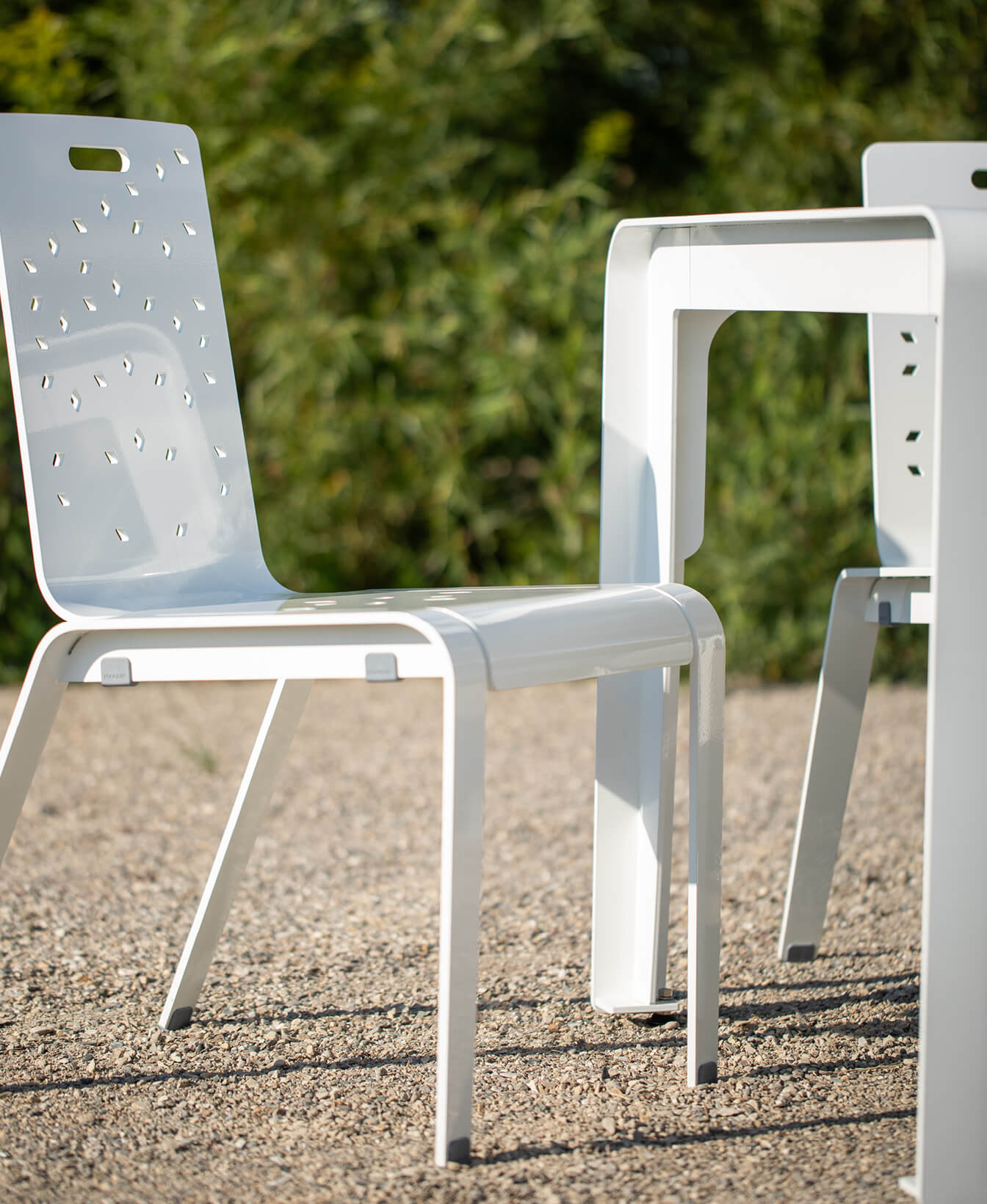An ALUM Café Chair featuring a white metal frame and perforated backrest sits beside a matching table outdoors on gravel, with greenery in the background.
