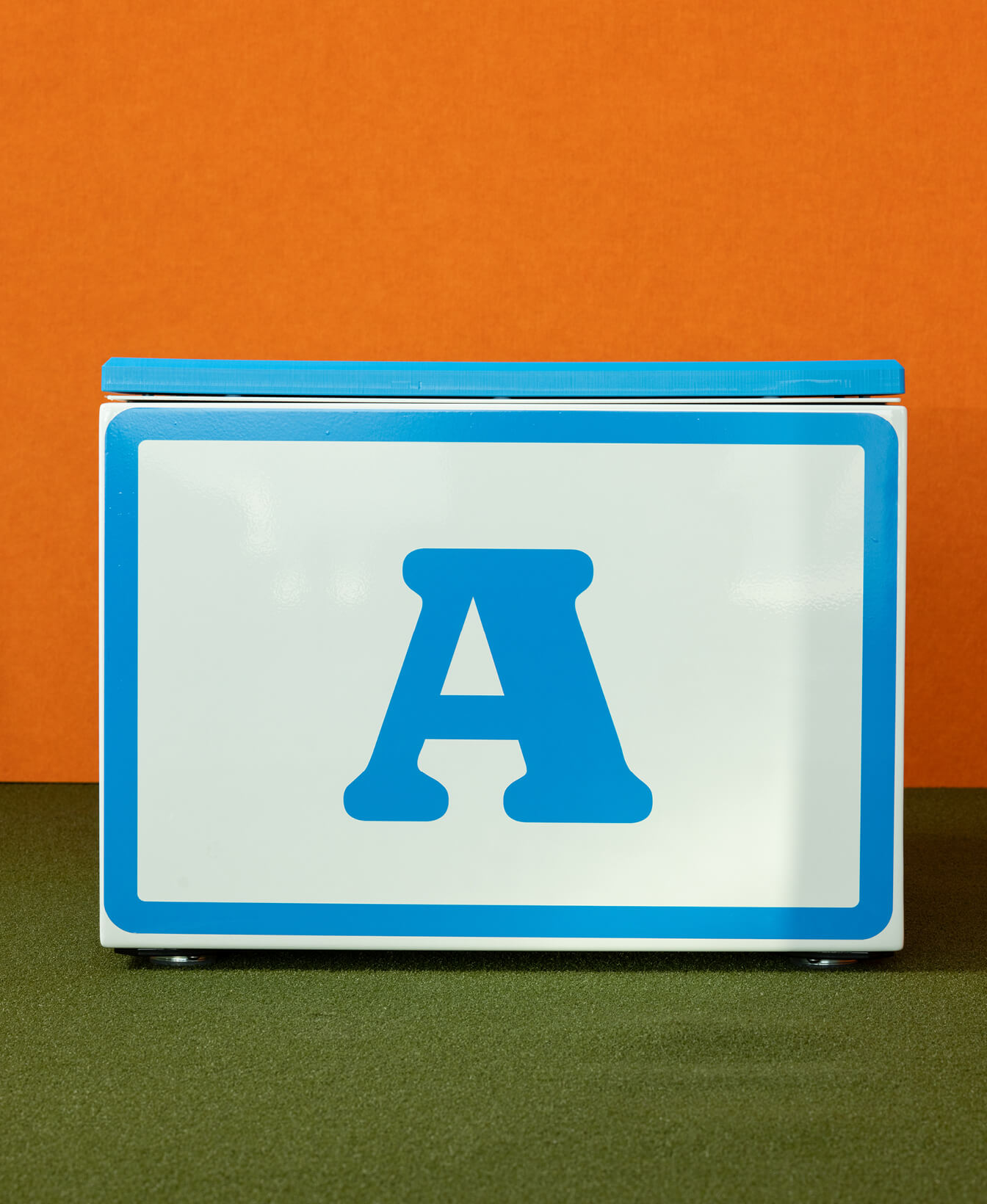 Une boîte Blocs Pixel avec un grand "A" bleu sur la façade blanche, une bordure bleue sur le couvercle, présentée sur un mur orange et un sol vert.