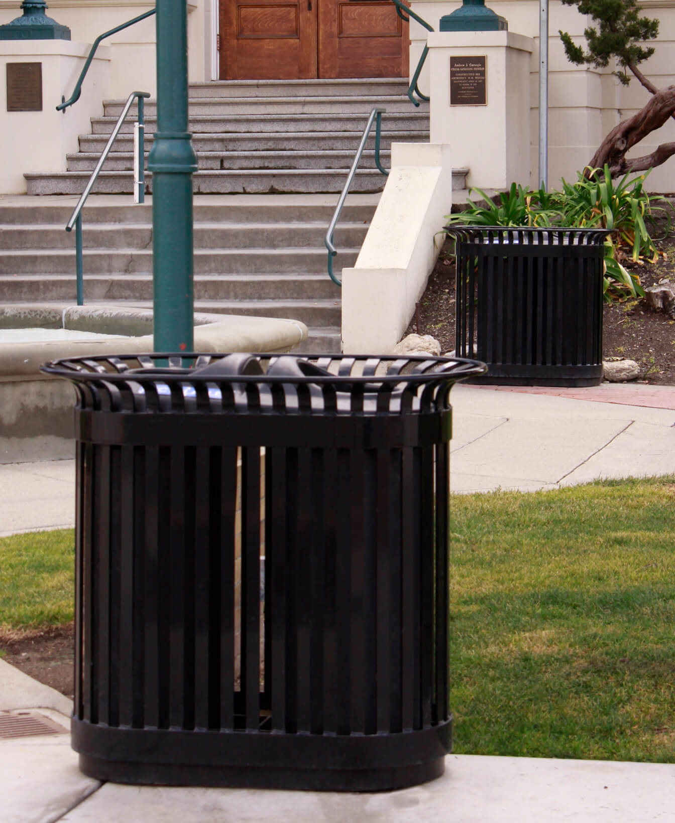 Un Contenant pour recyclage 200, une poubelle de recyclage en métal noir, se trouve sur le trottoir près d'un escalier et de l'entrée d'un bâtiment, avec une autre poubelle visible à l'arrière-plan.