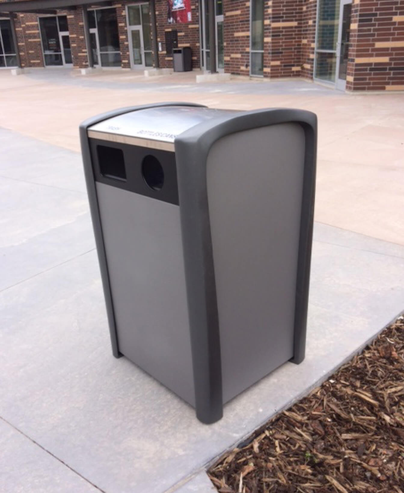 Un Contenant pour recyclage 1400, une poubelle extérieure grise pour le recyclage et les déchets, est placé sur une allée en béton à côté d'un bâtiment aux murs de briques et aux portes vitrées.