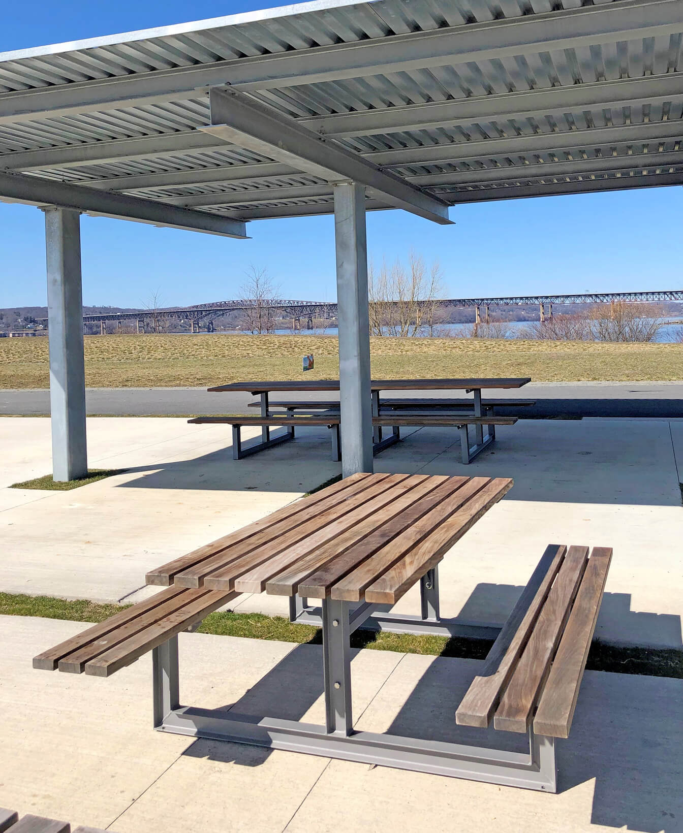Table et sièges groupés 210, avec des cadres en métal et des plateaux en bois, sont installés sur une surface en béton dans un parc extérieur avec un pont et de l'eau visible en arrière-plan.