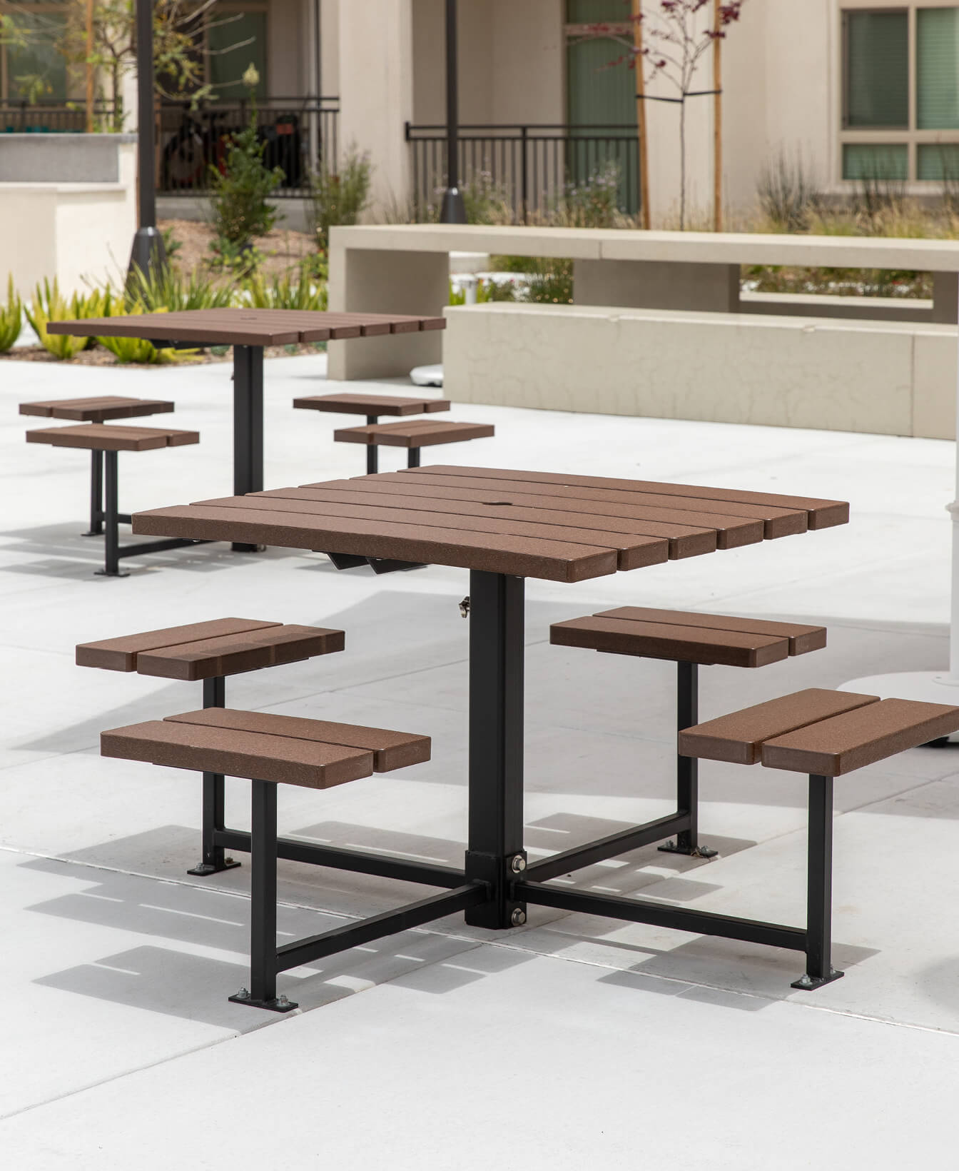 The 400 Cluster Seating features brown wooden tables with attached benches, perfect for outdoor patios surrounded by modern architecture and landscaping.
