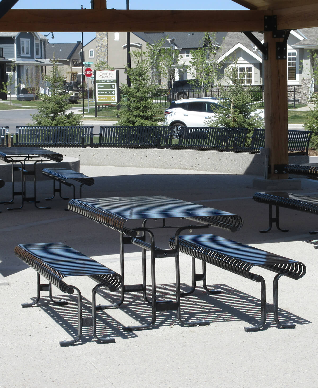 Espace extérieur couvert présentant la Table et sièges 510 avec des tables et des bancs en métal sur une surface en béton ; un quartier résidentiel et une voiture garée sont visibles à l'arrière-plan.