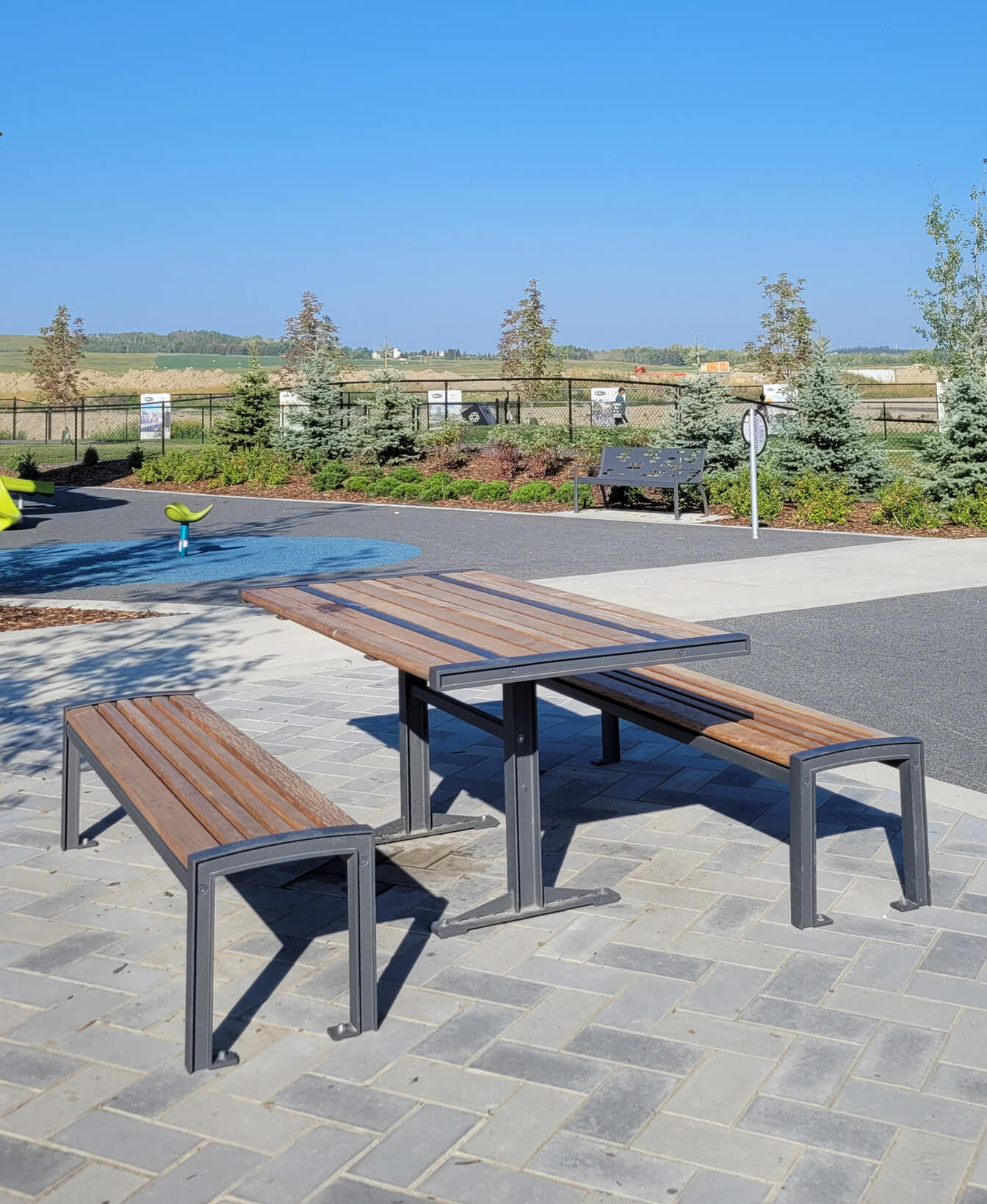 The 720 Table and Benches, made of wood, are placed on a paved area in an outdoor park surrounded by trees and open landscape beneath a clear blue sky.