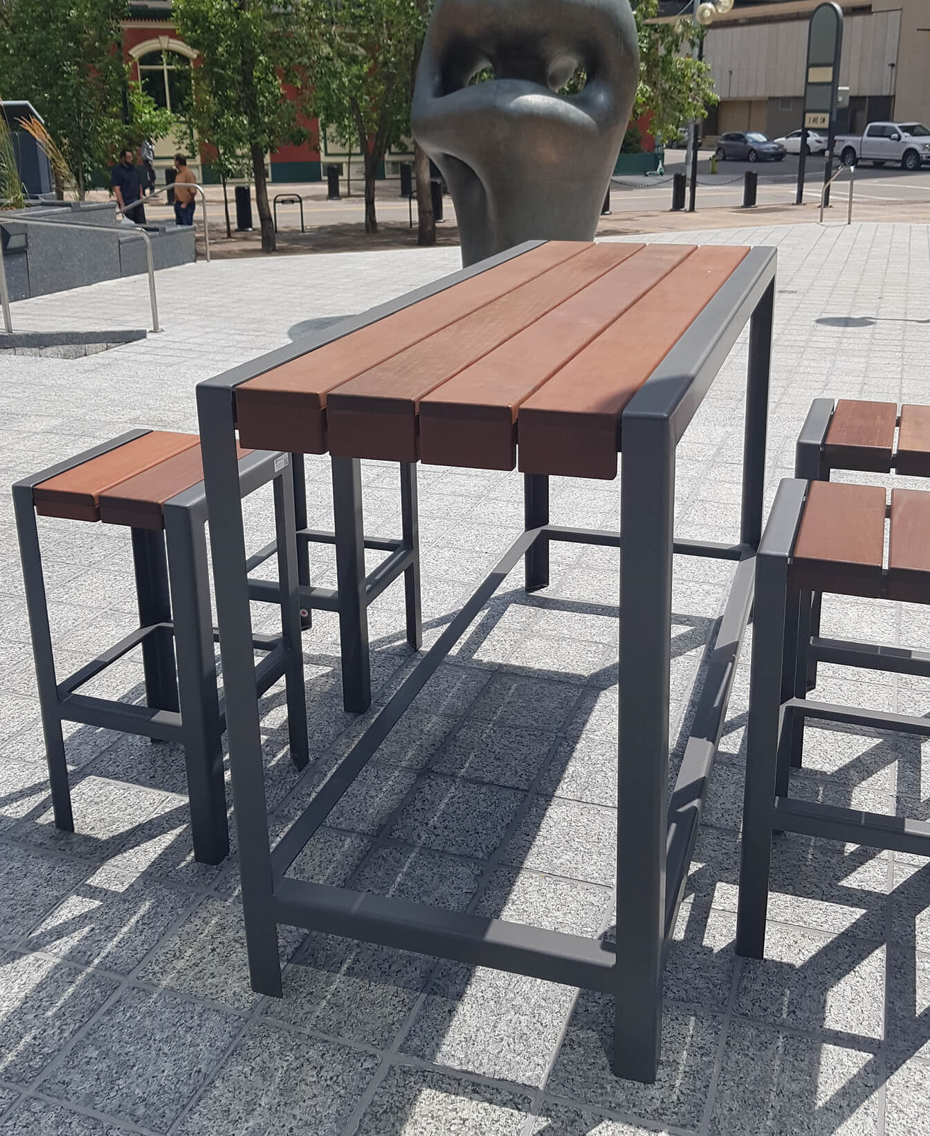 The 1050 Bar Height Tables, featuring metal and wood construction with matching benches, are placed on a paved public area surrounded by trees and a large abstract sculpture in the background.