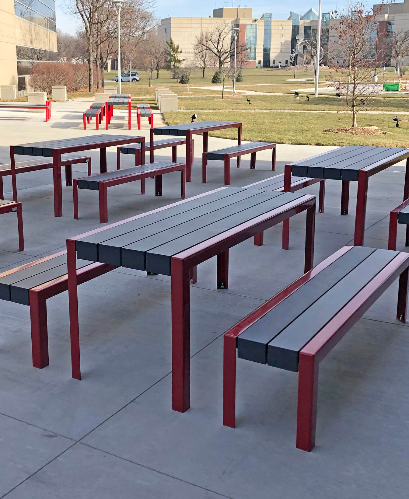 Several 1050 Dining Tables with matching benches are set up on a concrete patio outdoors, surrounded by grass and modern buildings in the background.