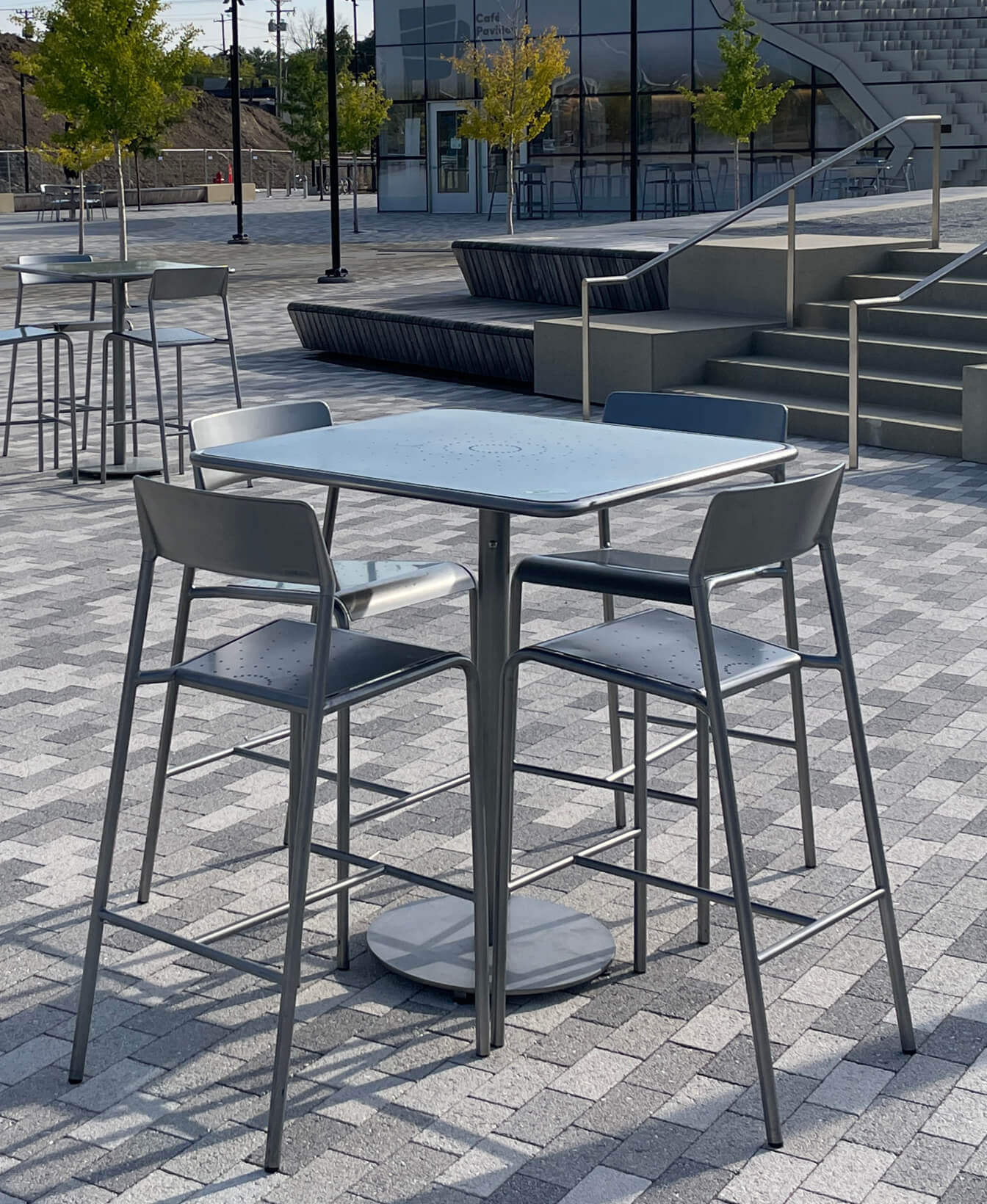 A Foro Bar Height Table with four matching high chairs sits on a gray patterned outdoor patio; additional tables and trees appear in the background.
