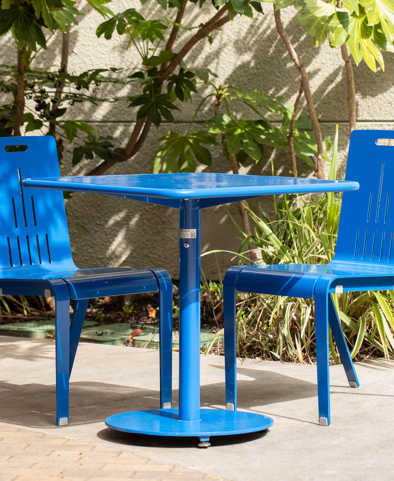 The 1100 Cluster Seating, featuring a blue metal square table and two matching blue chairs, is arranged outdoors on a patio with green plants in the background.
