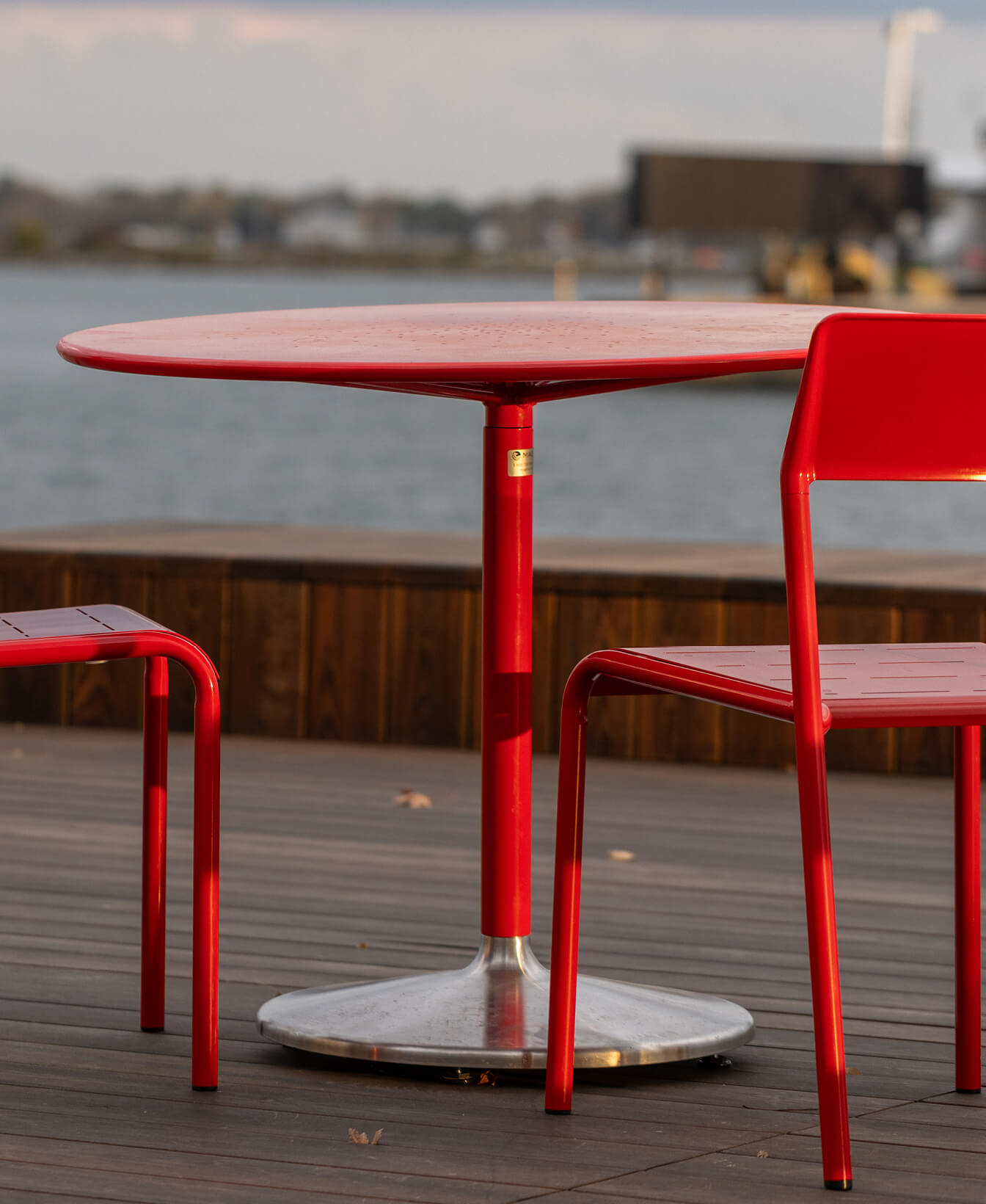 Une Table à café Battery rouge et deux chaises assorties sont disposées sur une terrasse en bois surplombant l'eau, avec une scène industrielle en arrière-plan.