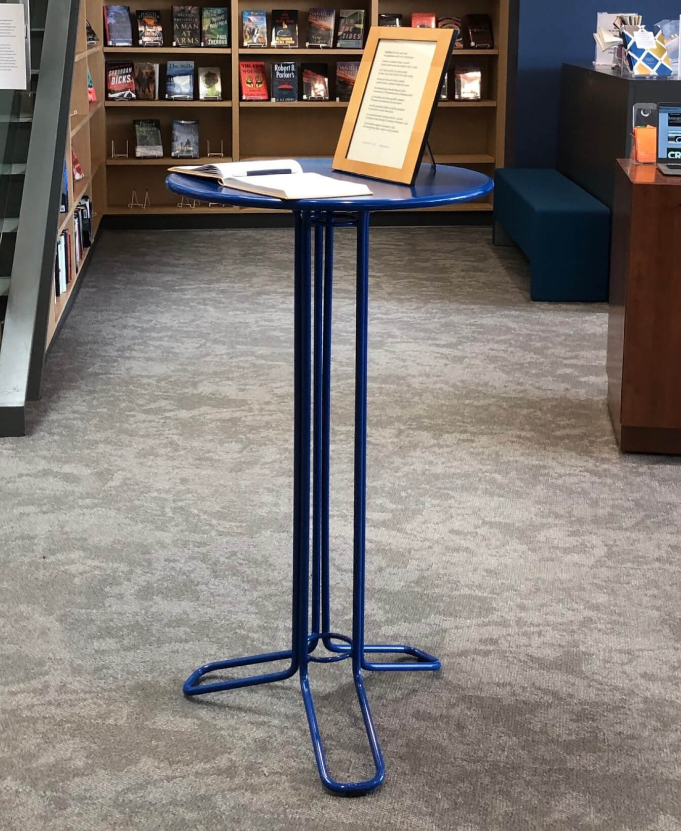 The Kontur Bar Height Table, featuring a small blue round top and thin metal legs, holds an open book and a framed document in a library with bookshelves and a carpeted floor.