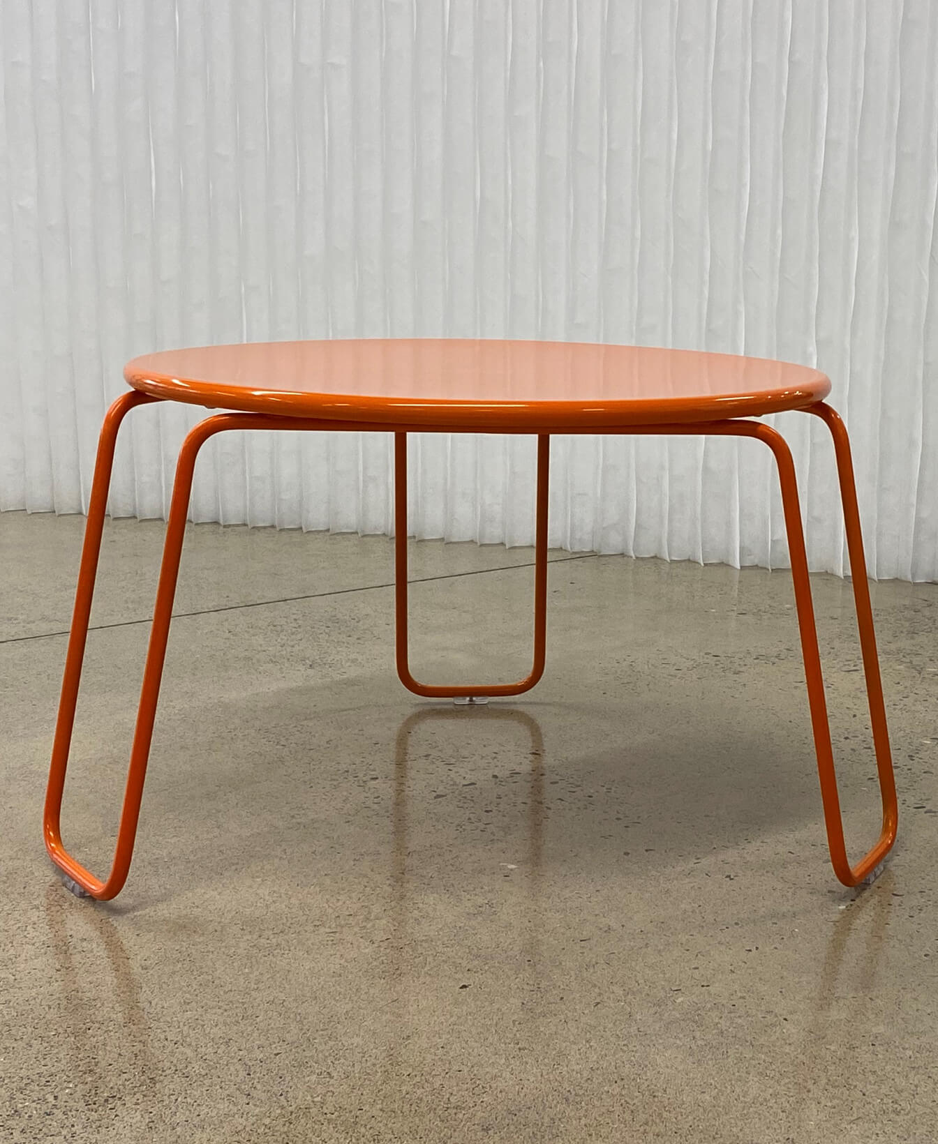 La Table basse Kontur, une table ronde en métal orange avec trois pieds angulaires, est posée sur un sol en béton poli devant un rideau blanc texturé.