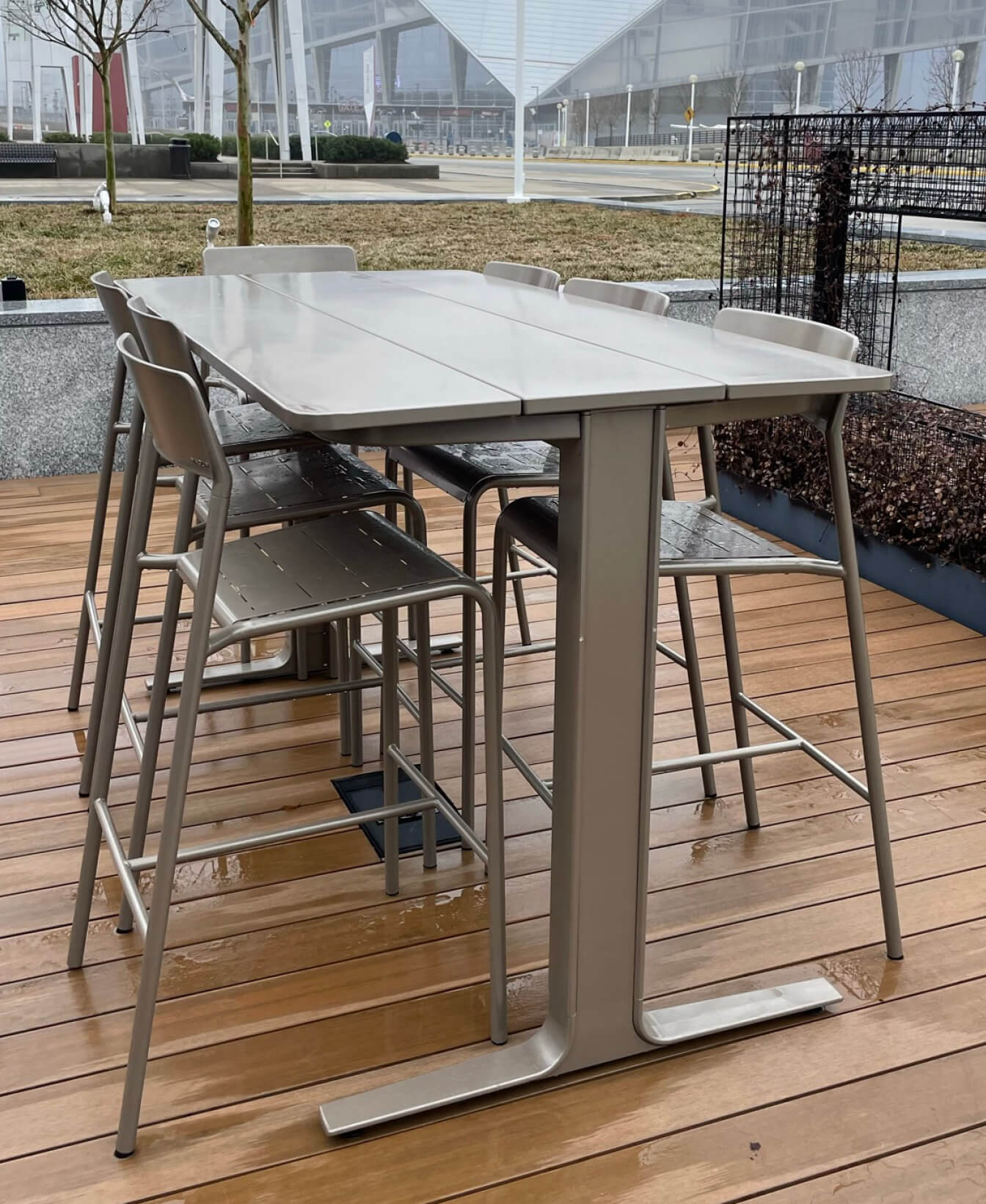 The Ancora Bar Height Table, paired with six matching stools, is set on a wooden deck outdoors with buildings and landscaped surroundings in the background.