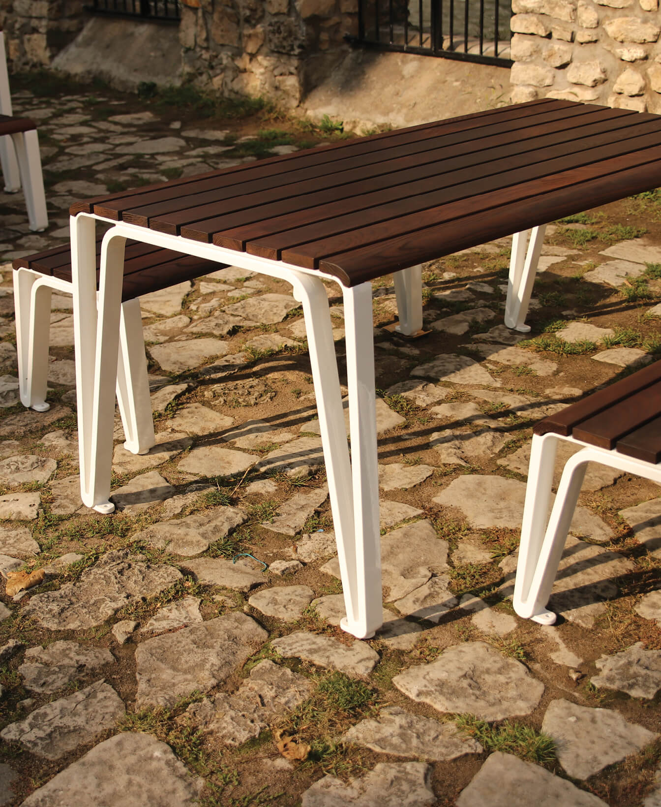 The Iconic Table, featuring a wooden top and white metal legs, stands on a cobblestone surface with matching benches.
