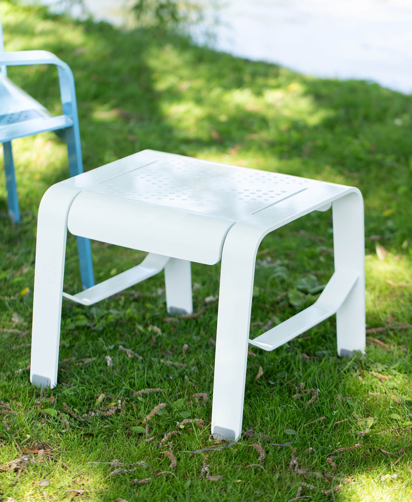The ALUM Lounge Table, a white metal table with a perforated top, is set on grass outdoors with part of a blue chair visible nearby.