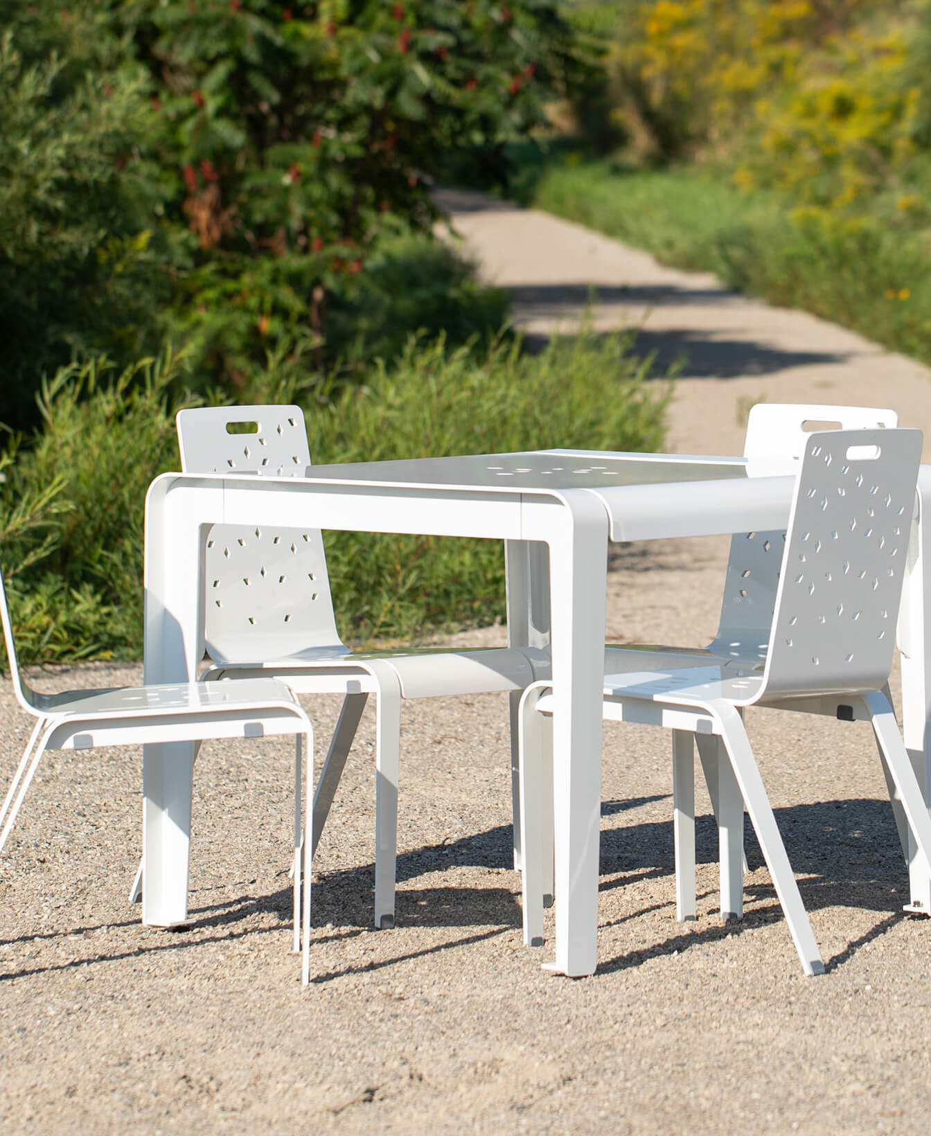 An ALUM Café Table with four matching chairs is set outdoors on a gravel path, surrounded by greenery and a dirt trail in the background.