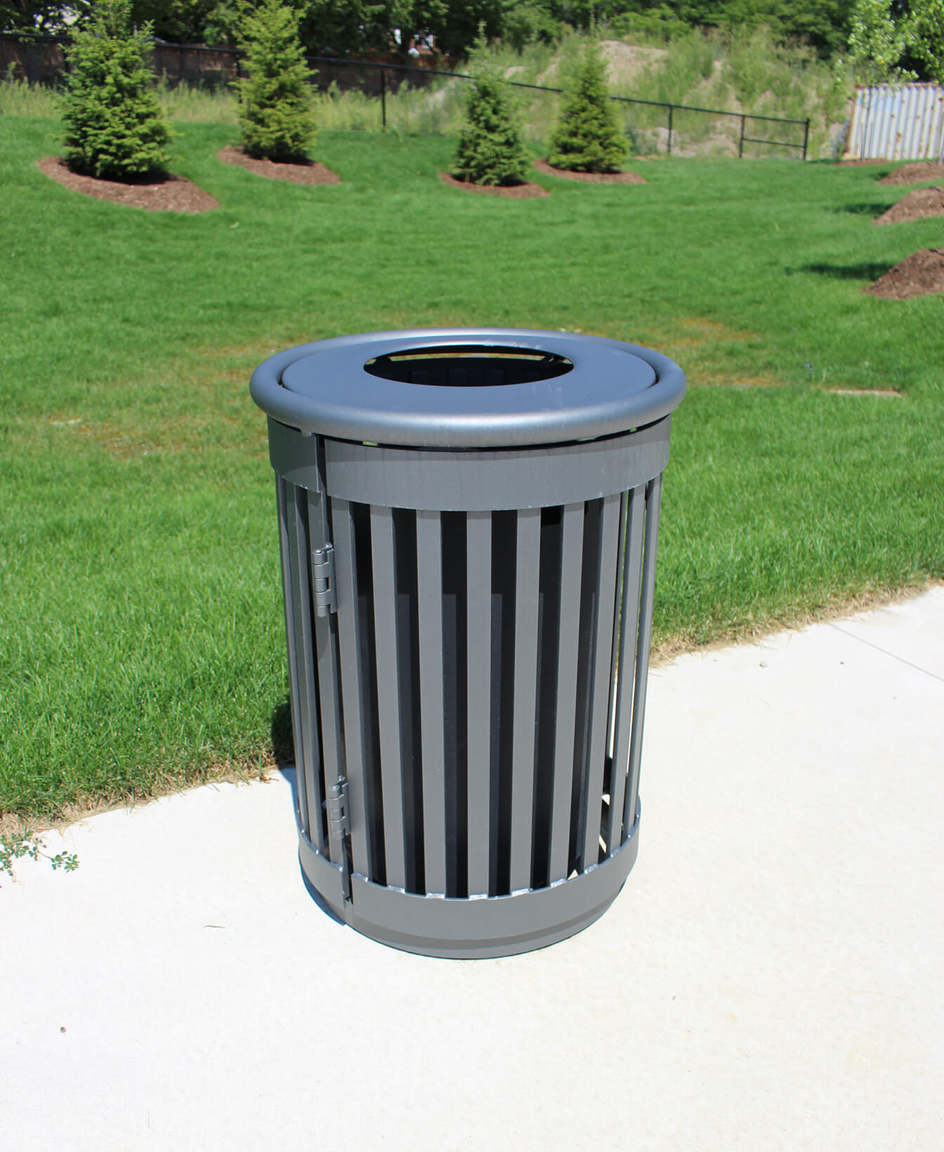 The 250 Trash Container, featuring a gray finish and vertical slats, is placed on a concrete path in a grassy park area with small trees in the background.