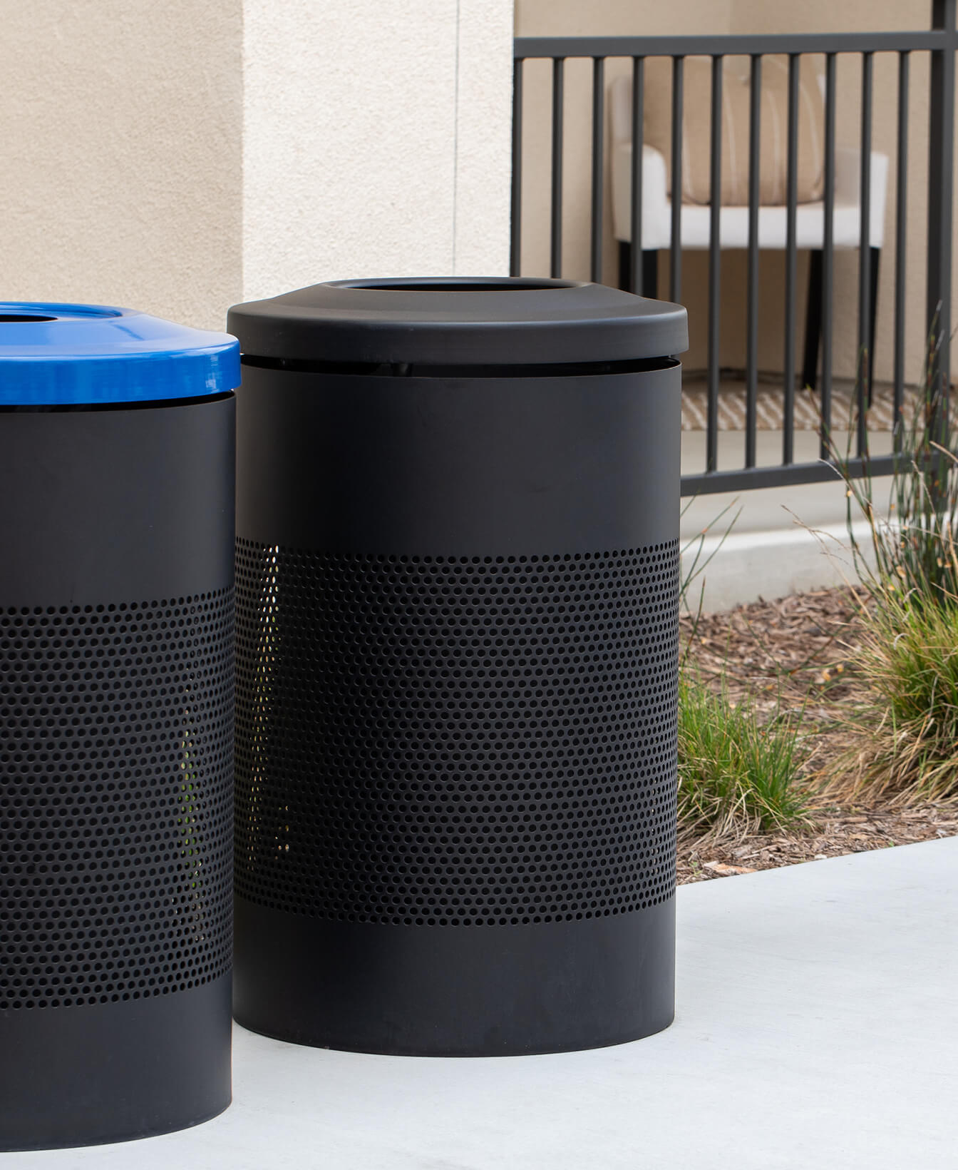 Two 600 Trash Container bins, one black with a blue lid and one all black, are placed on a concrete surface near a building and metal railing.
