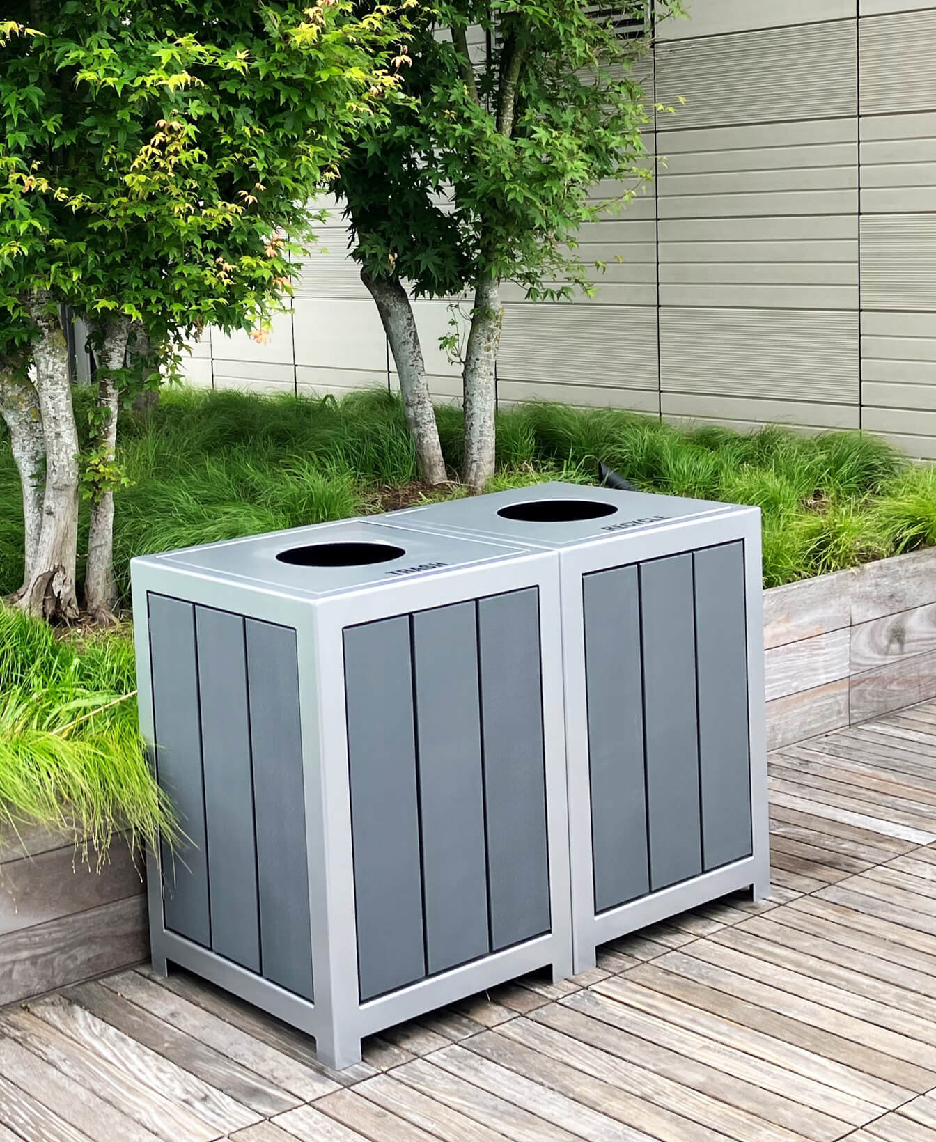 Deux Contenant à déchets 1050 gray sont placés sur une terrasse en bois près d'une jardinière avec de l'herbe verte et des arbres.