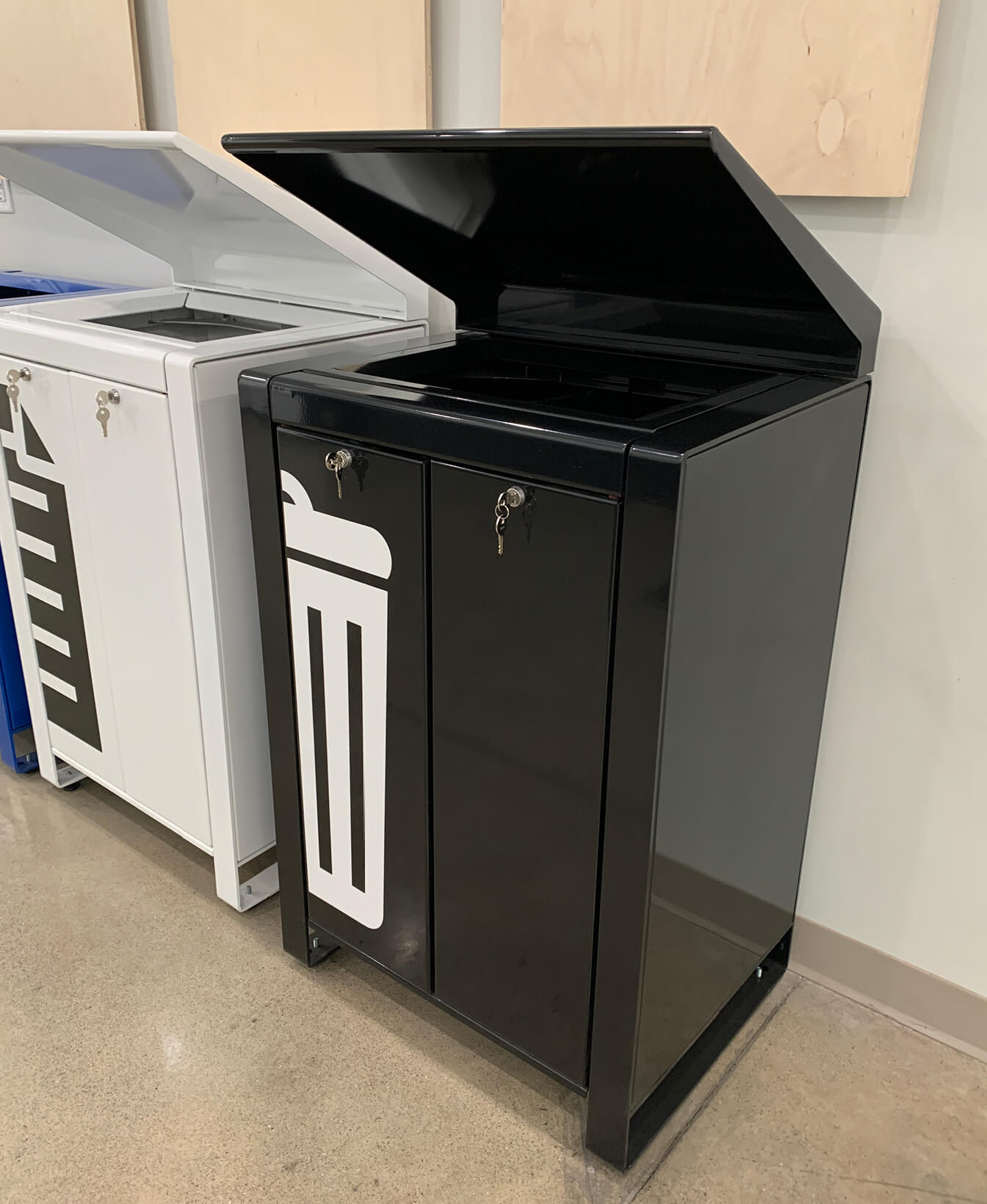 A black Lexicon Trash Container with a locking door and open top lid stands beside a similar white bin on a polished indoor floor.
