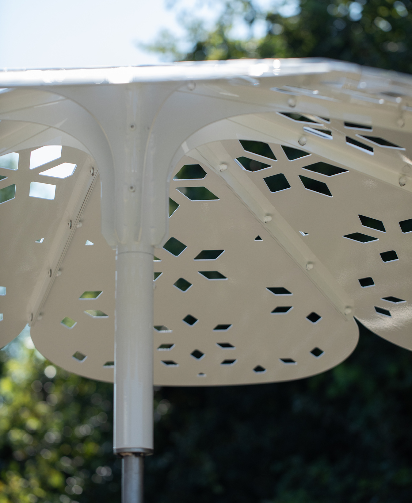 Gros plan sur le parasol ROMA, une structure métallique blanche avec des motifs géométriques découpés, sur un fond flou d'arbres et de ciel.