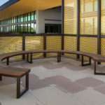 Les Bancs Ogden, bancs en bois courbés, sont placés sur une terrasse extérieure carrelée, entourée de panneaux métalliques avec des découpes circulaires, et se trouvent à côté d'un bâtiment doté de grandes baies vitrées.