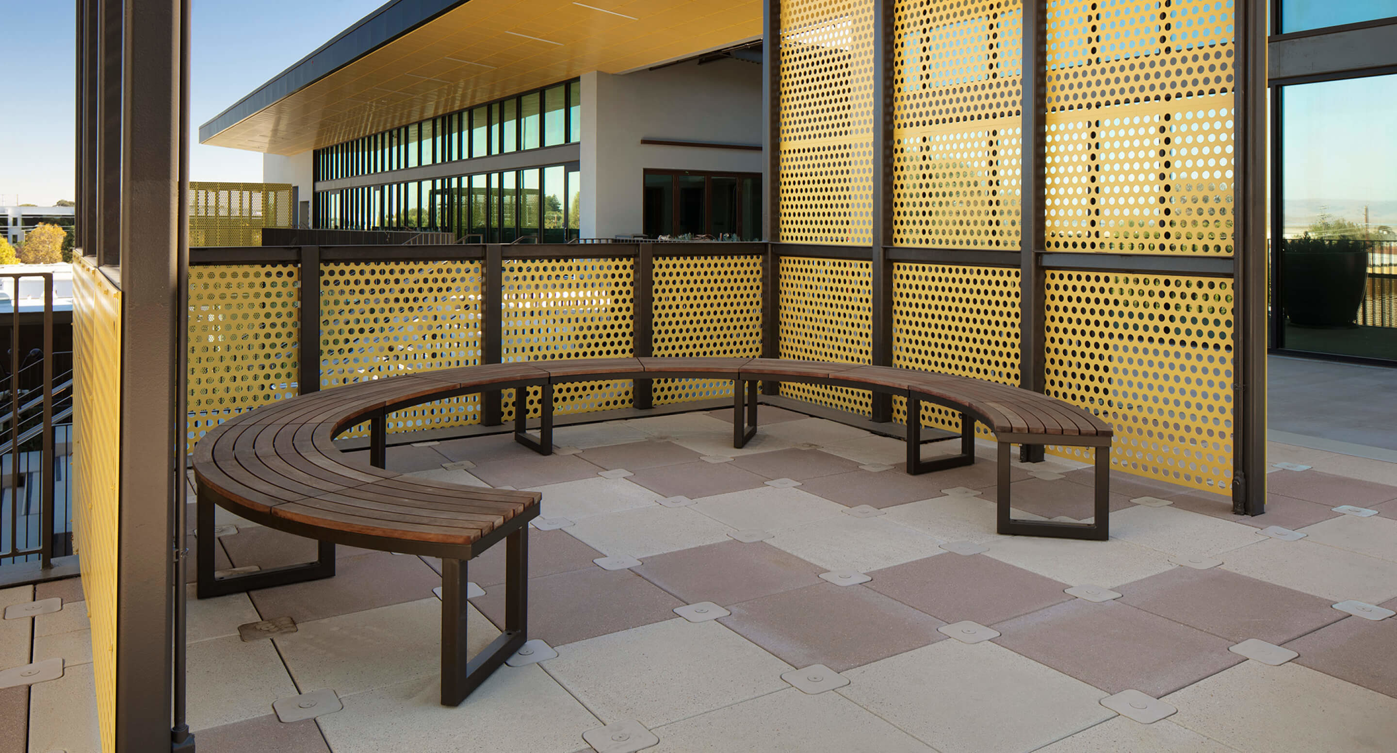 Les Bancs Ogden, bancs en bois courbés, sont placés sur une terrasse extérieure carrelée, entourée de panneaux métalliques avec des découpes circulaires, et se trouvent à côté d'un bâtiment doté de grandes baies vitrées.