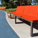 Le Bancs Ogden, doté d'un siège orange vif et d'une armature métallique, est placé sur un chemin en béton à côté d'une surface caoutchoutée bleue dans un parc entouré d'herbe et d'arbres.