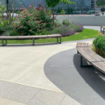 Bancs Ogden Des bancs en bois courbés bordent une allée pavée dans un parc paysager avec des buissons verts, des plantes à fleurs et un bâtiment en arrière-plan.