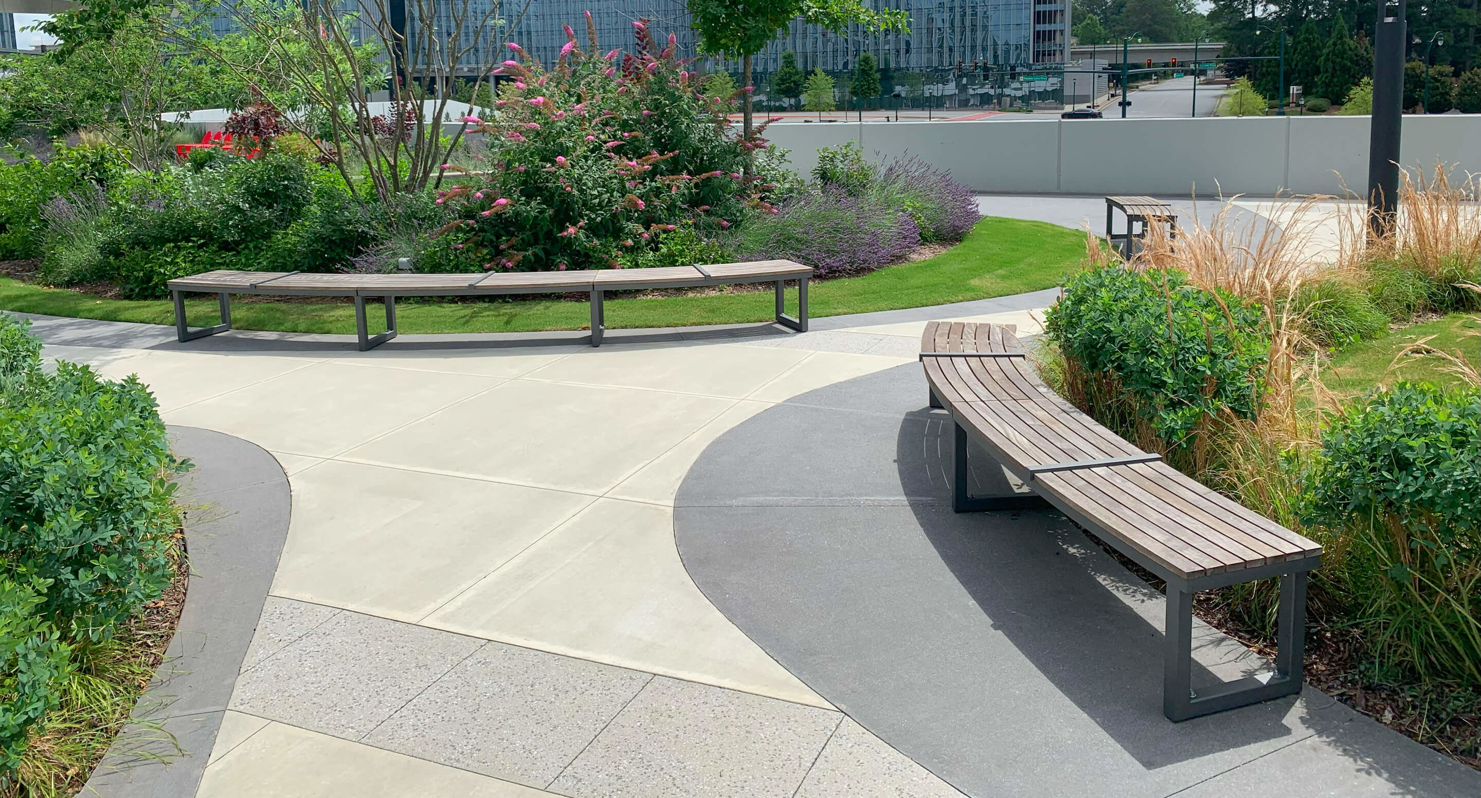 Bancs Ogden Des bancs en bois courbés bordent une allée pavée dans un parc paysager avec des buissons verts, des plantes à fleurs et un bâtiment en arrière-plan.