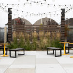 Un patio extérieur avec des bancs Bancs Ogden, des tables rondes jaunes, des guirlandes lumineuses et de hautes herbes près d'une clôture en lattes de bois, niché entre deux bâtiments blancs modernes.
