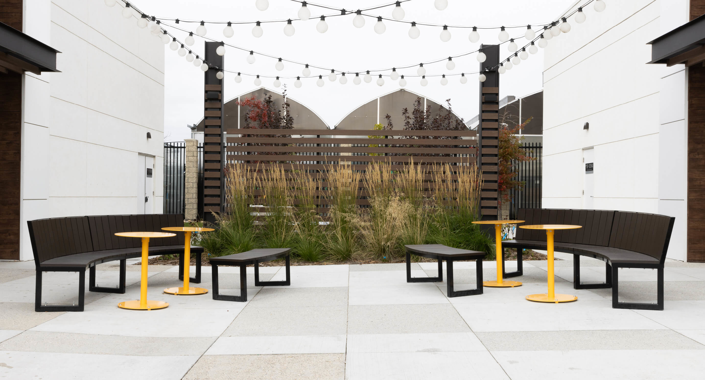 Un patio extérieur avec des bancs Bancs Ogden, des tables rondes jaunes, des guirlandes lumineuses et de hautes herbes près d'une clôture en lattes de bois, niché entre deux bâtiments blancs modernes.