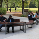 People sit on curved wooden benches near a sign for Messay Hall in a park-like campus setting, with trees and grass in the background.