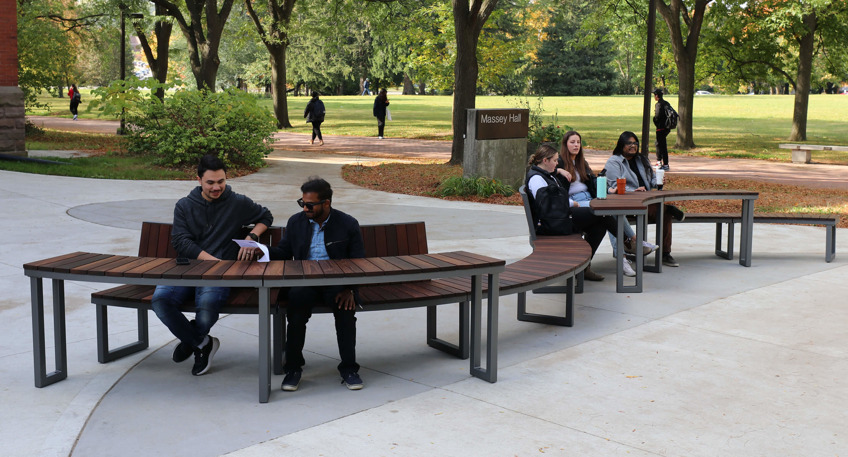 maglin_ogden-modular-table_2 People sit on curved wooden benches near a sign for Messay Hall in a park-like campus setting, with trees and grass in the background.
