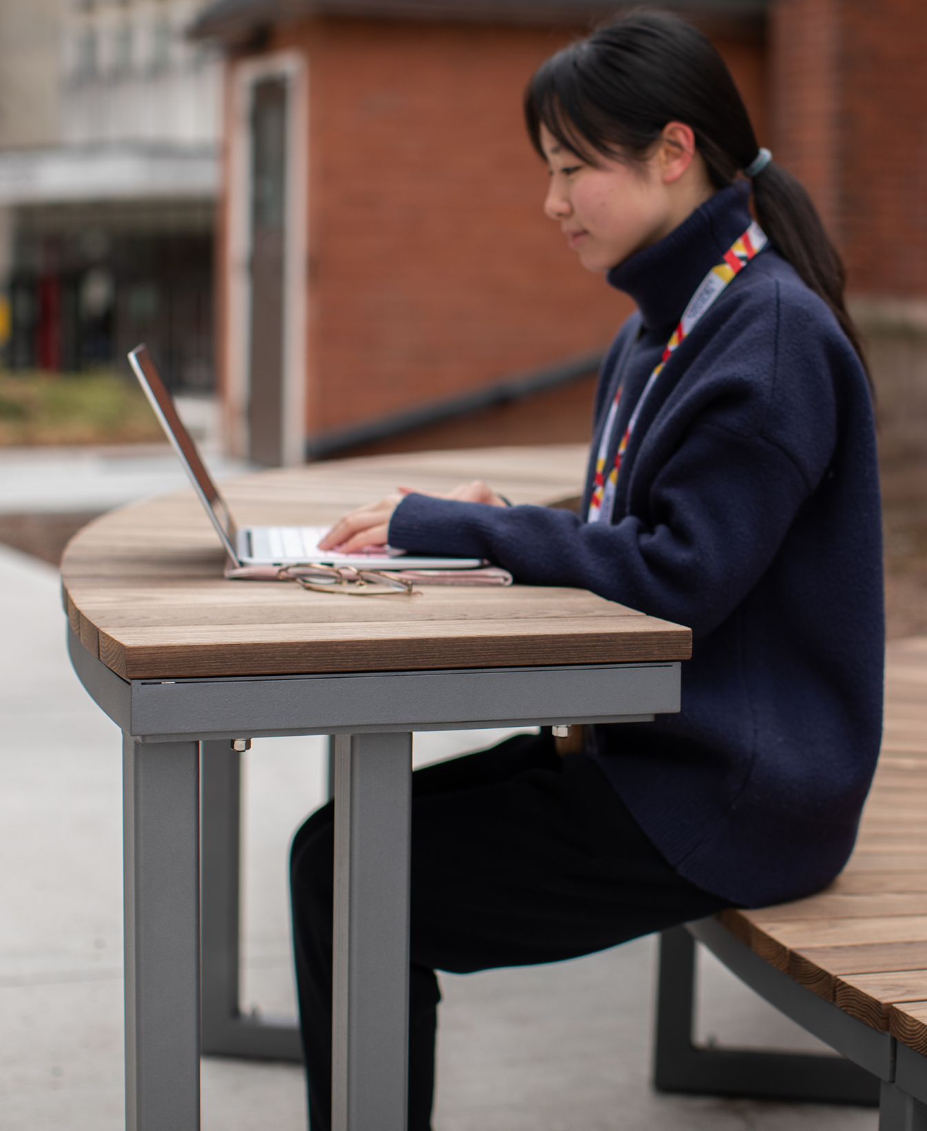 A person uses a laptop at the Ogden Modular Table outdoors, with eyeglasses resting on the table beside them.