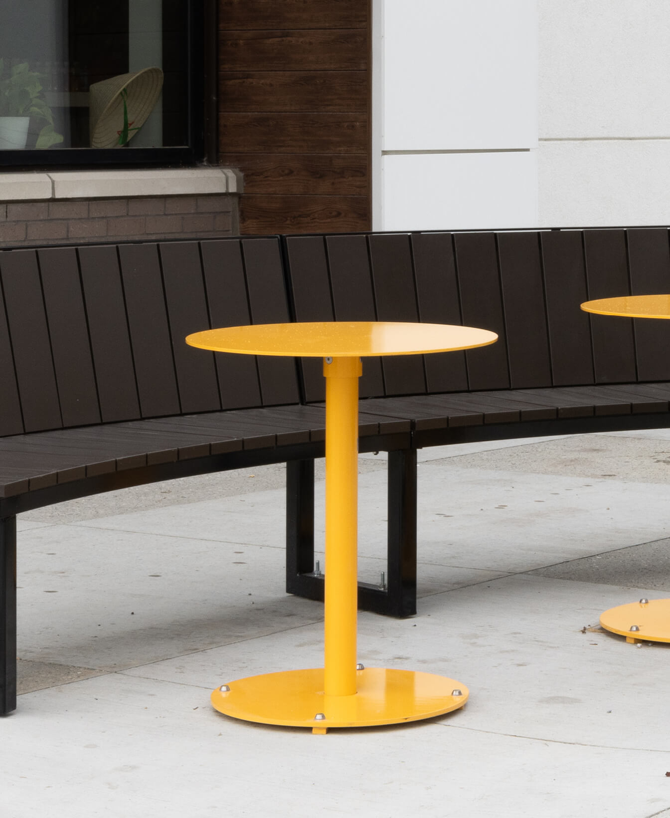 An Ogden Café Table, round and yellow, stands on a concrete sidewalk in front of a curved dark brown bench and a building with brick and white panel walls.