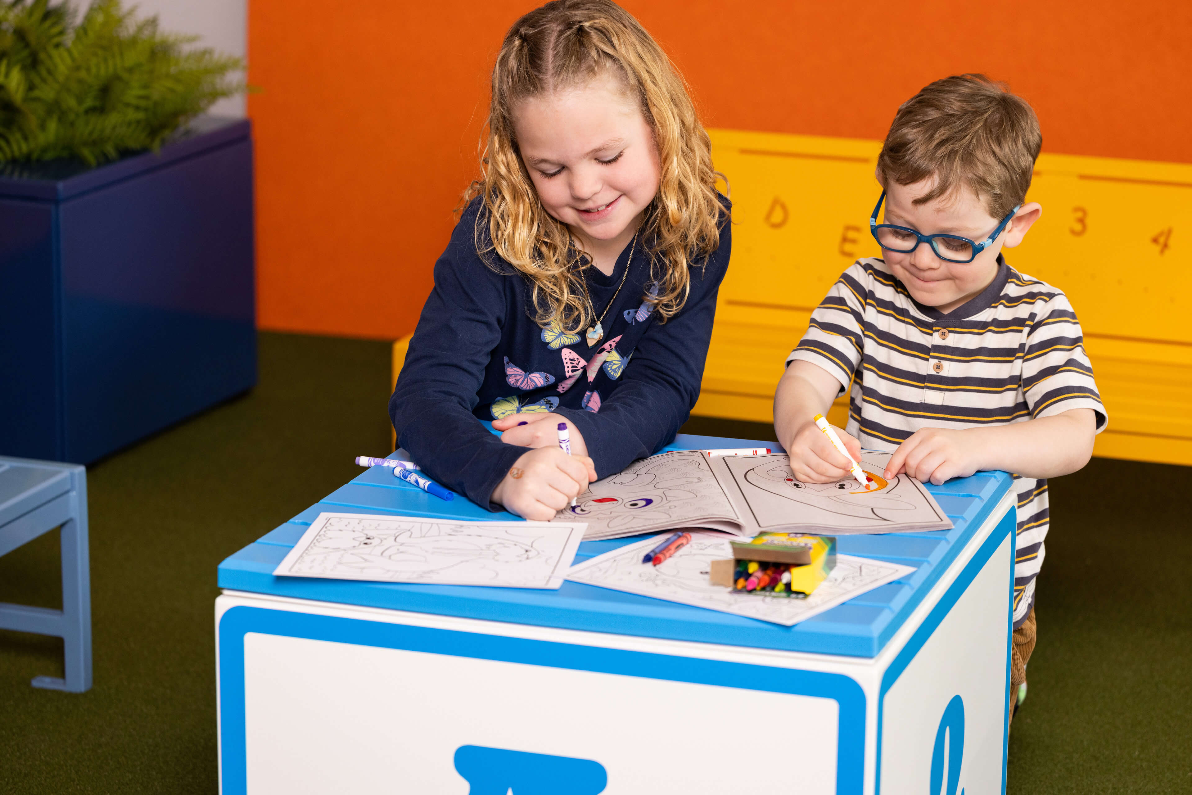 Two young children sit at a square table, coloring in books with crayons. A yellow bench and a blue chair are in the background.