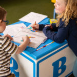 Deux enfants colorient dans des livres de coloriage sur un grand bloc décoré de la lettre 