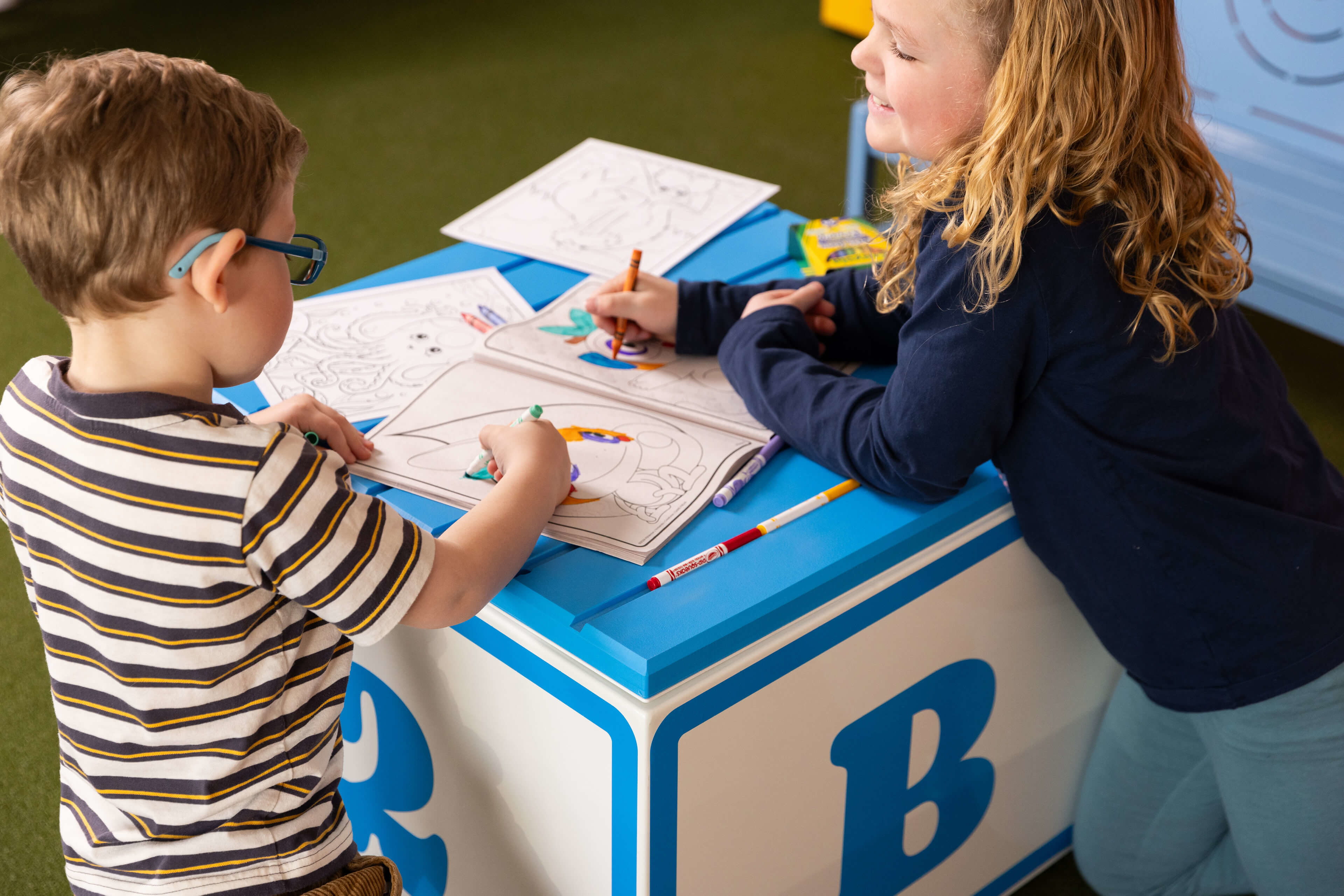 Deux enfants colorient dans des livres de coloriage sur un grand bloc décoré de la lettre 