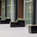 A row of black planter benches is arranged along the large glass windows of a modern building, with trees and a chair visible in the background.